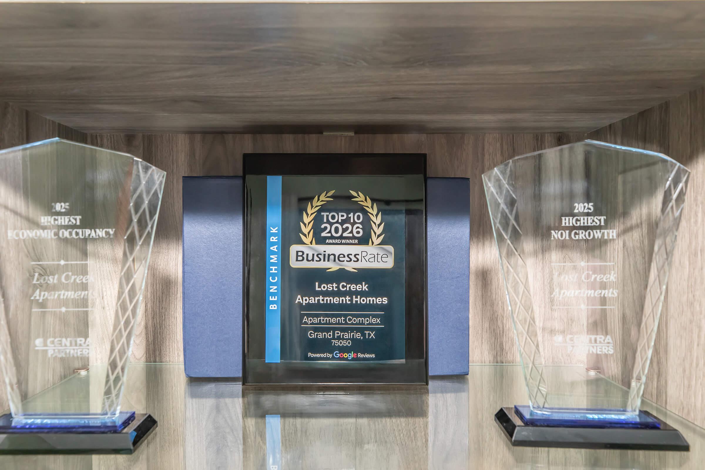 A display case containing three awards. The center award is a plaque recognizing "Lost Creek Apartment Homes" for being in the "Top 10 2026" in the BusinessRate category. The two flanking awards highlight achievements for “Highest Economic Occupancy” and “Highest NOI Growth” for the property located in Grand Prairie, TX.