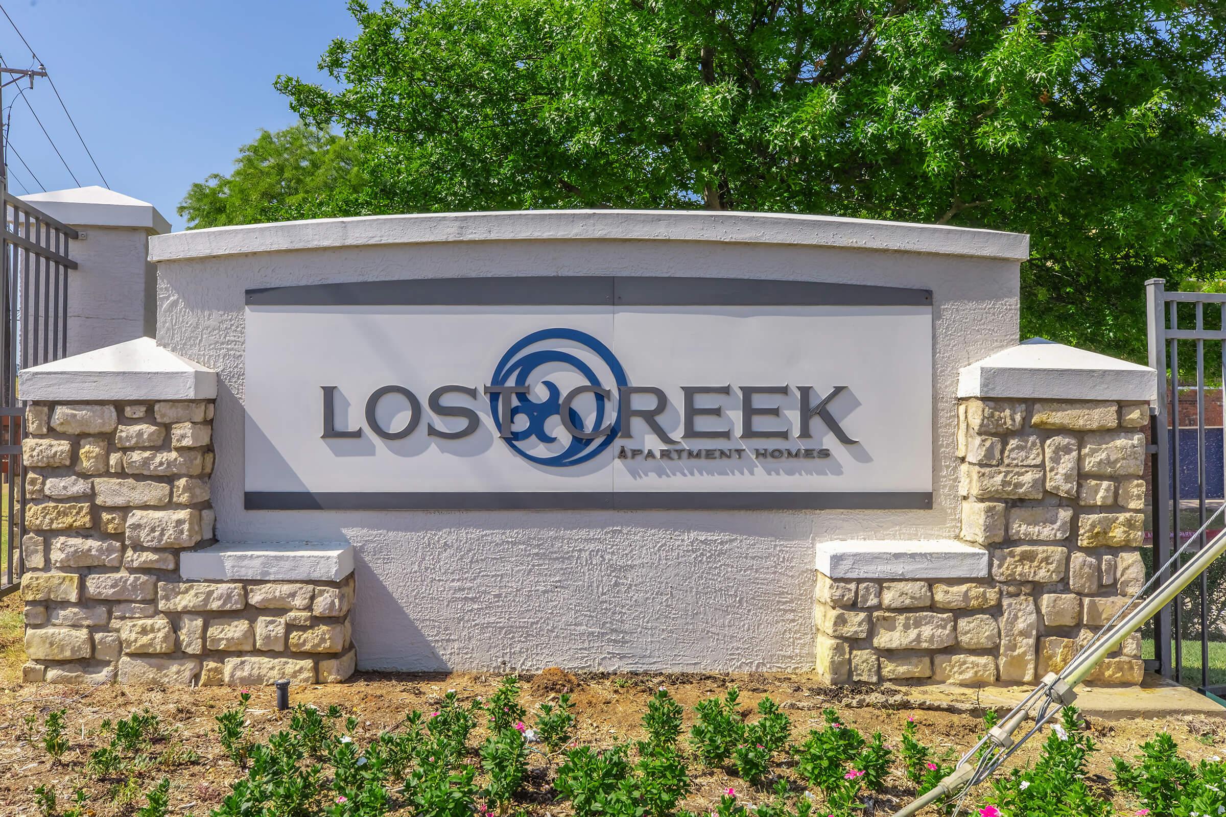Sign for Lost Creek Apartment Homes, featuring the name prominently displayed in blue letters with a stylized logo. The sign is bordered by a natural stone base and surrounded by greenery and flowers, set against a clear sky.