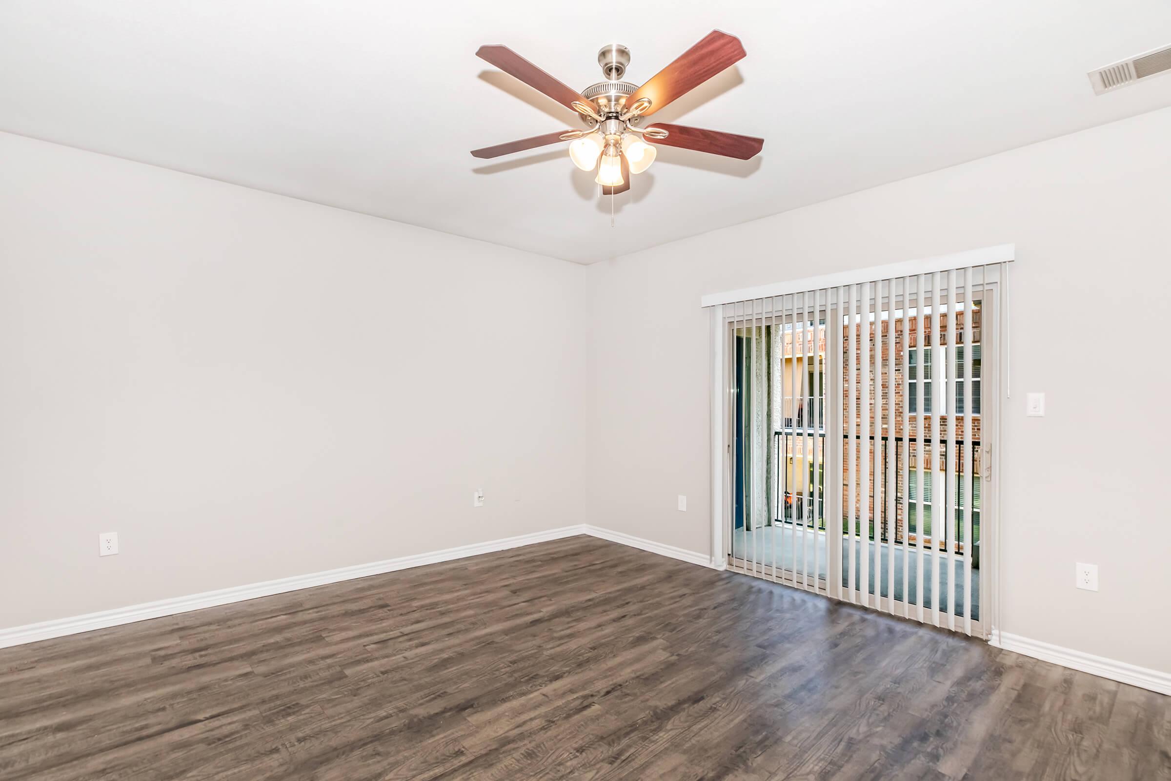 A spacious, empty room featuring a ceiling fan with wooden blades, light-colored walls, and a large sliding glass door leading to a balcony. The floor is a dark hardwood finish, contributing to a modern and airy atmosphere. Natural light enters through the door, enhancing the open feel of the space.