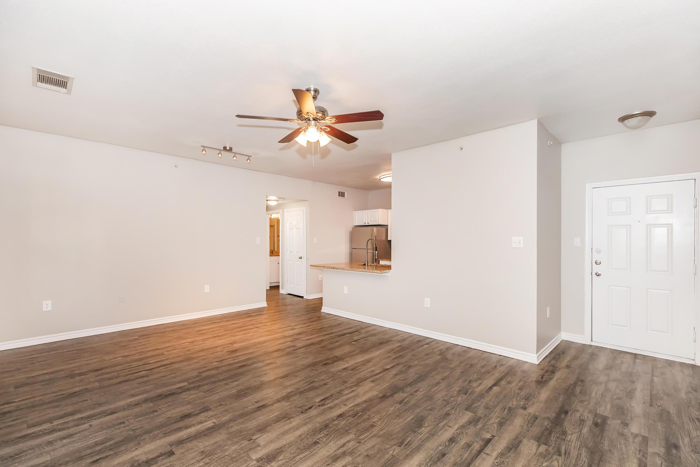 A spacious, empty living room featuring light-colored walls, a ceiling fan, and hardwood-style flooring. The open layout connects to a kitchen area with visible cabinetry. Light streams in from multiple entry points, highlighting the room's size and potential for furnishings. A front door is also visible.