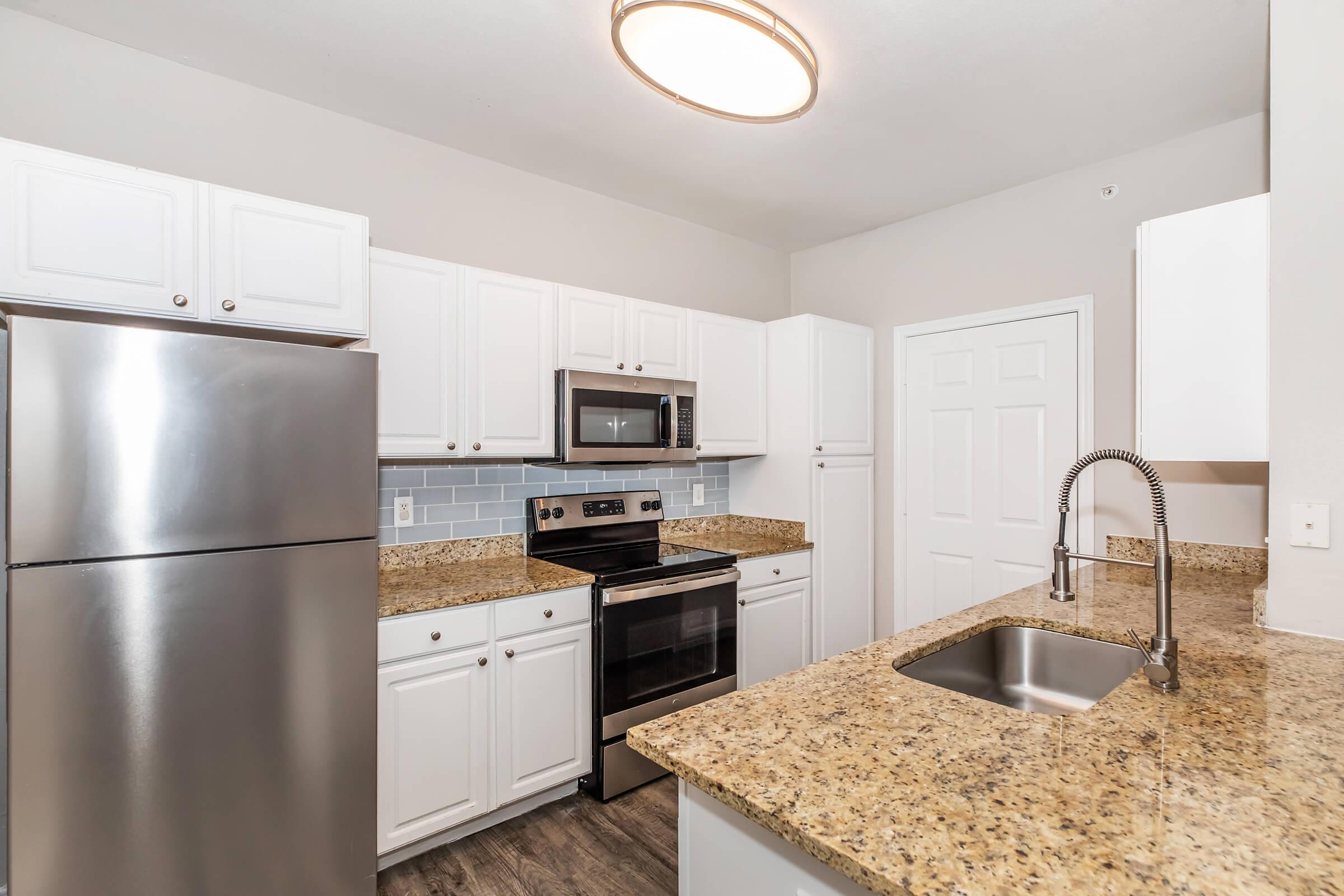 A modern kitchen featuring white cabinetry, stainless steel appliances including a refrigerator and microwave, a granite countertop, an undermount sink, and a subtle backsplash. The kitchen is well-lit with a ceiling light fixture and has a clean, contemporary design.