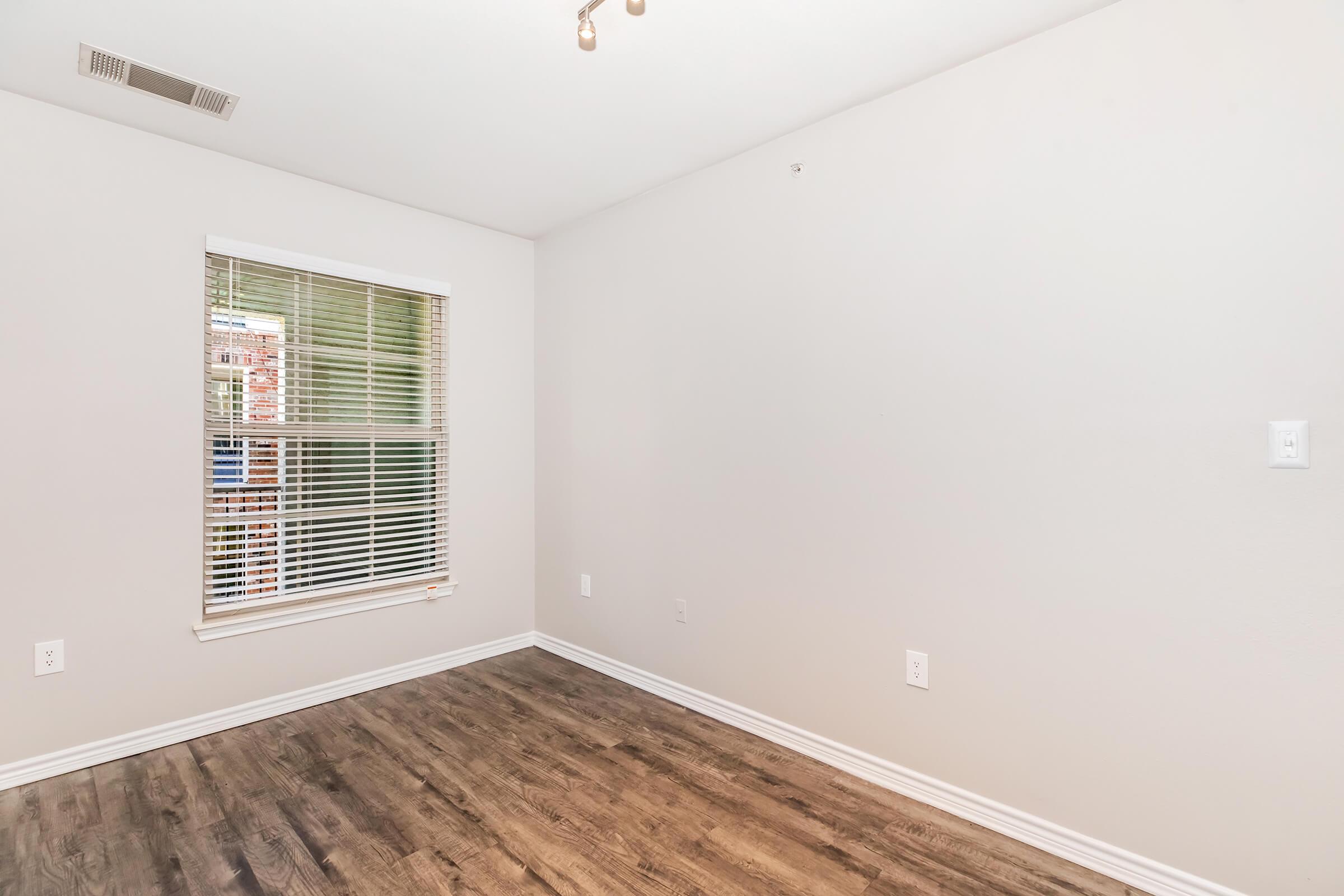 Empty room with light gray walls and dark wood laminate flooring. A window with white blinds provides natural light, and there are no furnishings or decorations visible. The space appears clean and ready for new occupants, with a ceiling light fixture and electrical outlets.