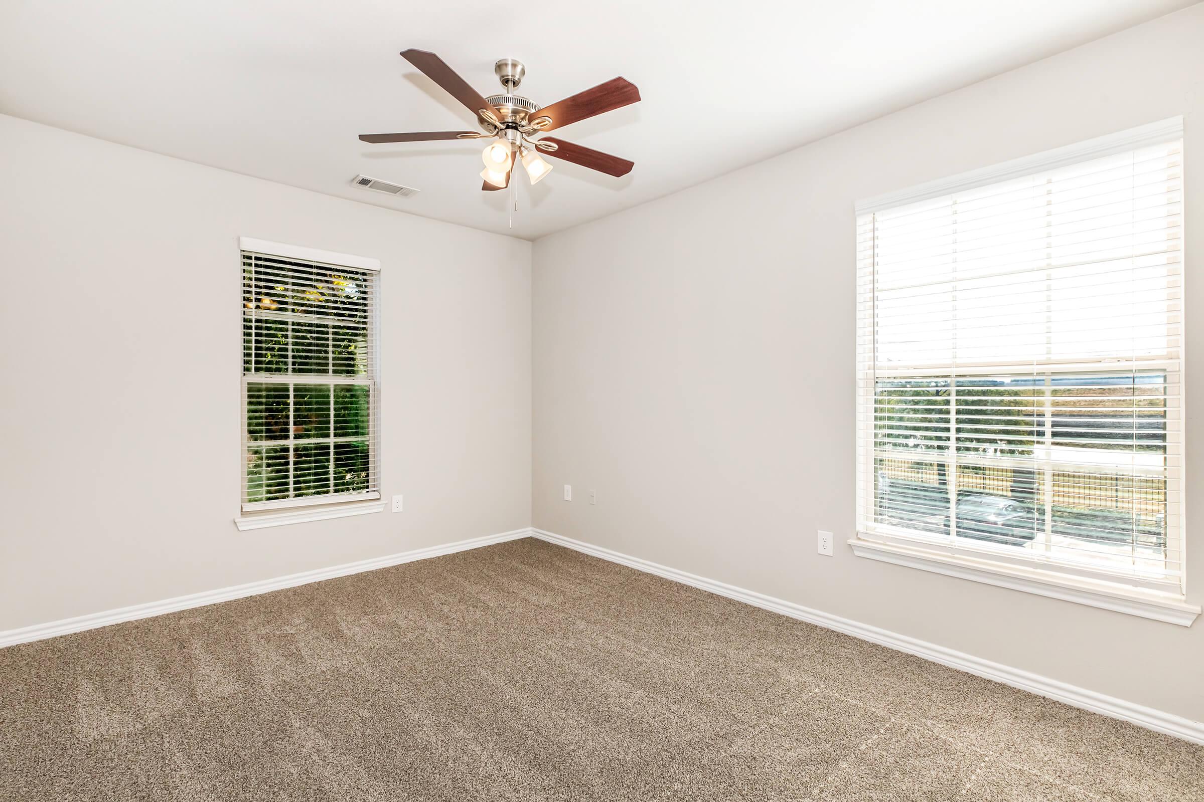Empty room featuring light-colored walls, a ceiling fan with wooden blades, and two windows with blinds. The floor is covered in beige carpet, and natural light streams in from the windows, illuminating the space.