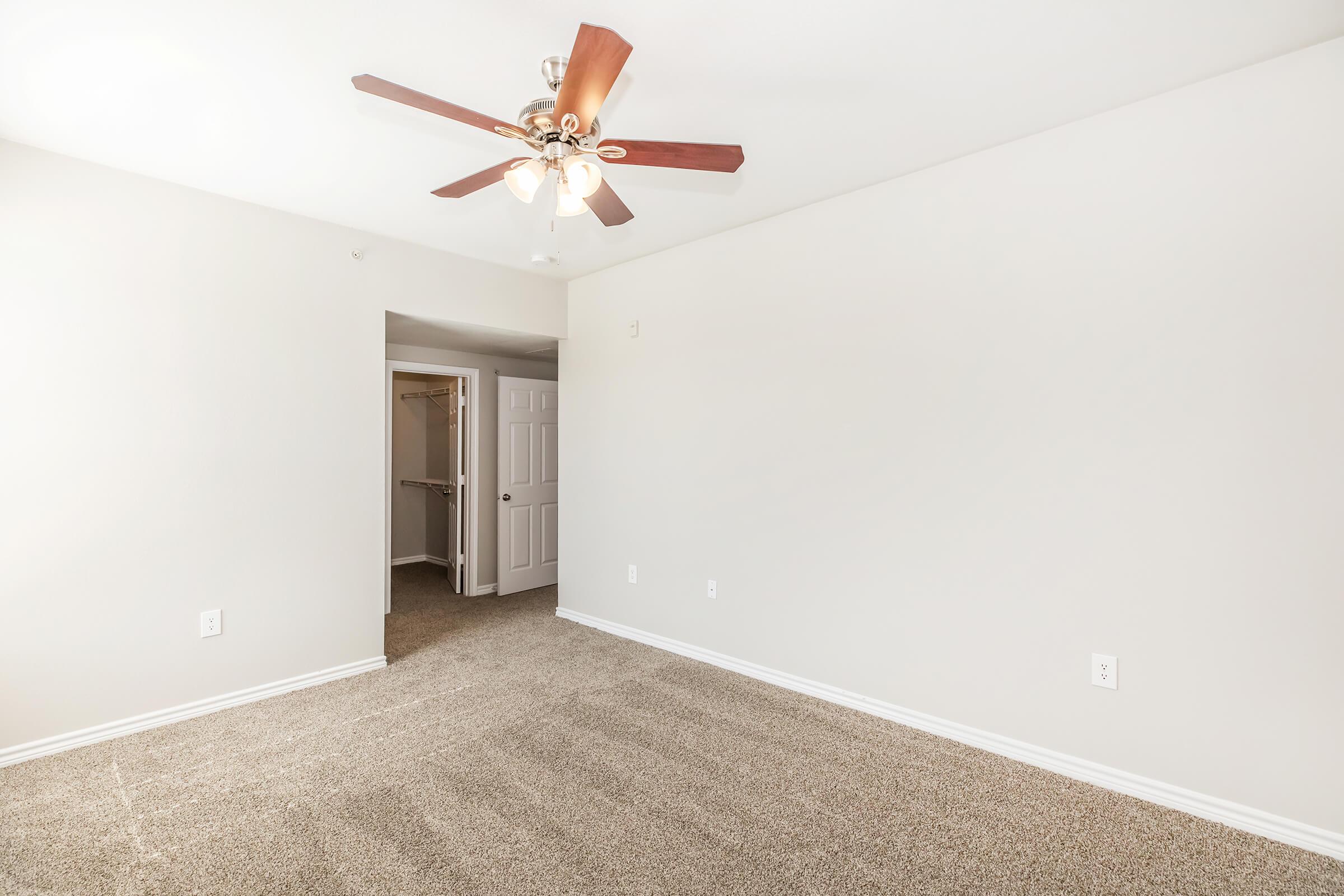 A spacious room with light beige walls and plush carpet. A ceiling fan with wooden blades is mounted in the center. A doorway is visible on the left, leading to a closet area. The overall atmosphere is bright and inviting, suitable for various uses like a bedroom or office.