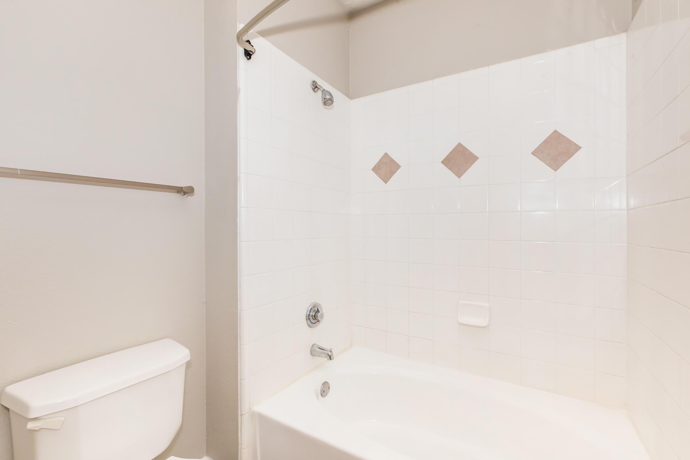A clean bathroom featuring a white bathtub with a showerhead and a white tiled wall adorned with decorative diamond-shaped tiles. A toilet sits nearby, and a towel bar is mounted on the wall. The overall aesthetic is bright and simple, with neutral colors.