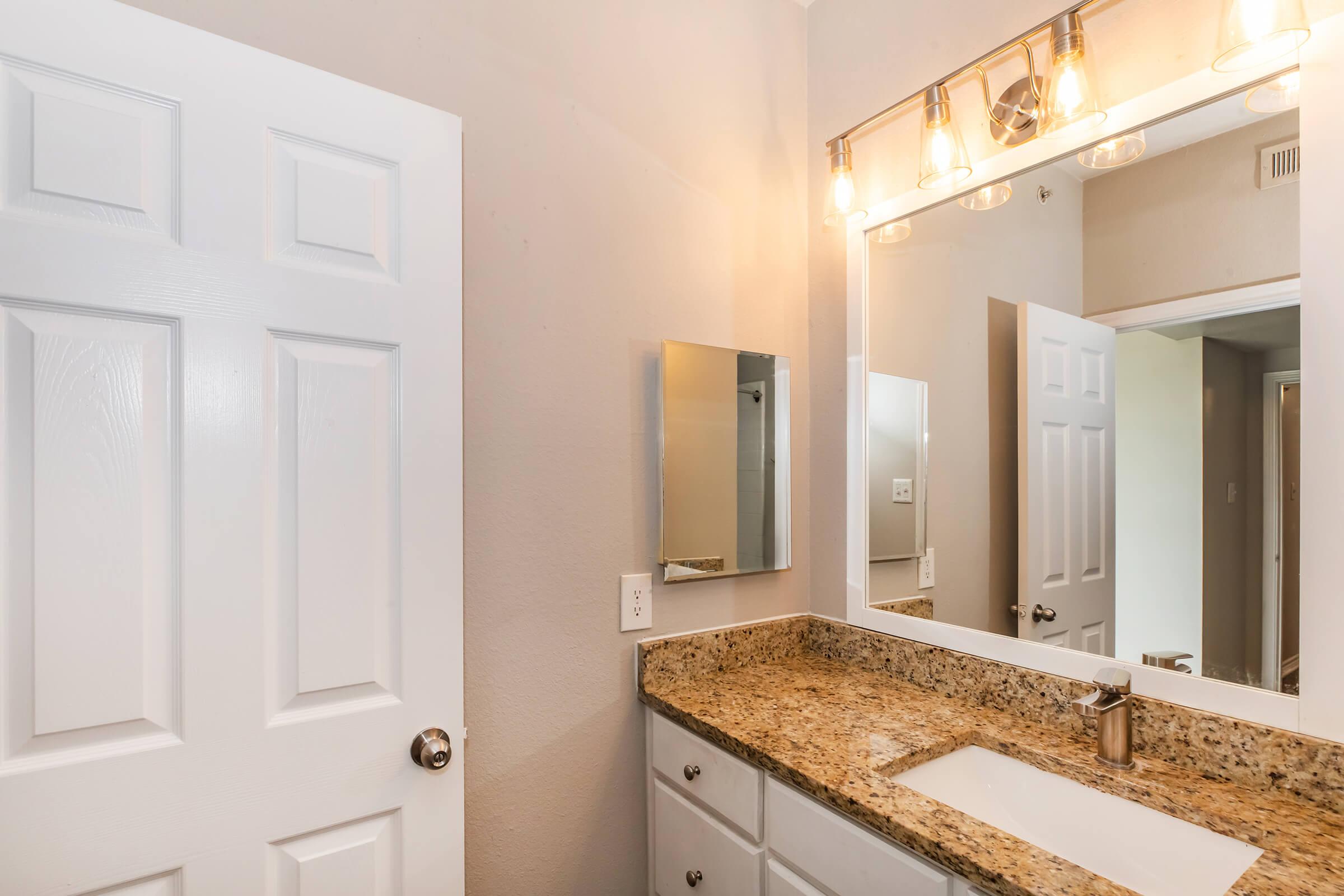 A well-lit bathroom featuring a white door, a large mirror above a granite countertop with a sink. A wall-mounted mirror is to the left, and there are light fixtures above the mirror casting a warm glow. The walls are painted in a light gray color, contributing to a modern and clean aesthetic.
