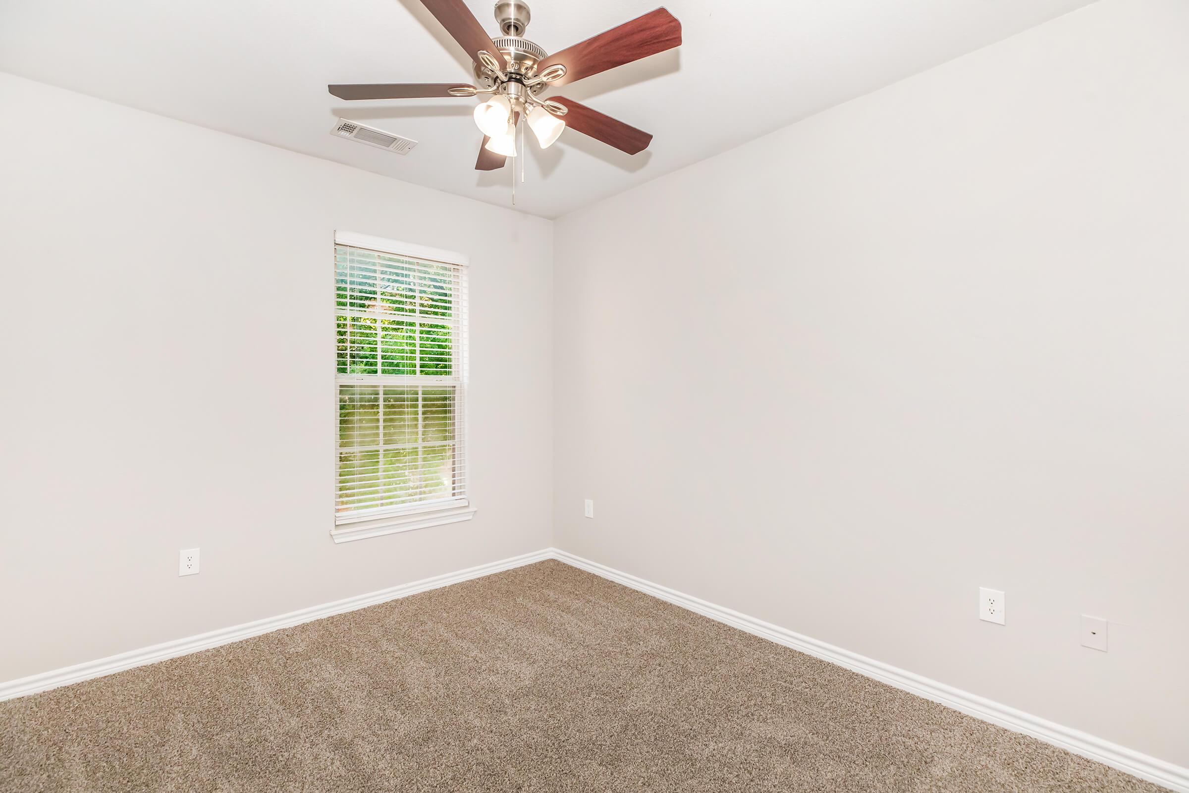 A well-lit empty room with light beige walls and a ceiling fan with wooden blades. It features a window with white blinds that lets in natural light, and the floor is covered with plush brown carpet. The room is unfurnished, providing a blank canvas for decoration.