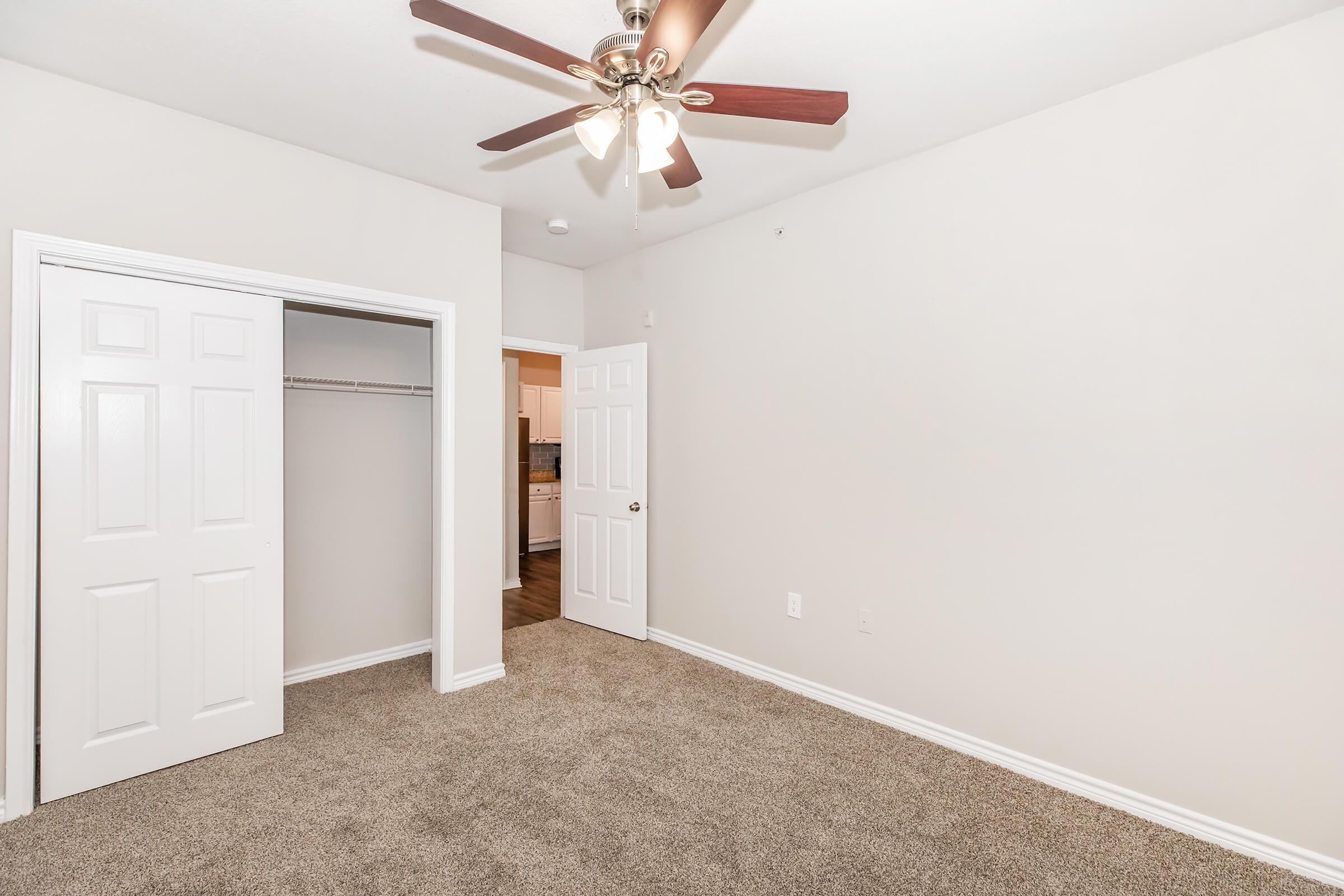 A view of a spacious, empty bedroom featuring light beige walls, a ceiling fan with three bulbs, and a plush carpet. There's a doorway with a white closet to the left and an open door leading to another room in the background. The space is well-lit and ready for furniture arrangement.