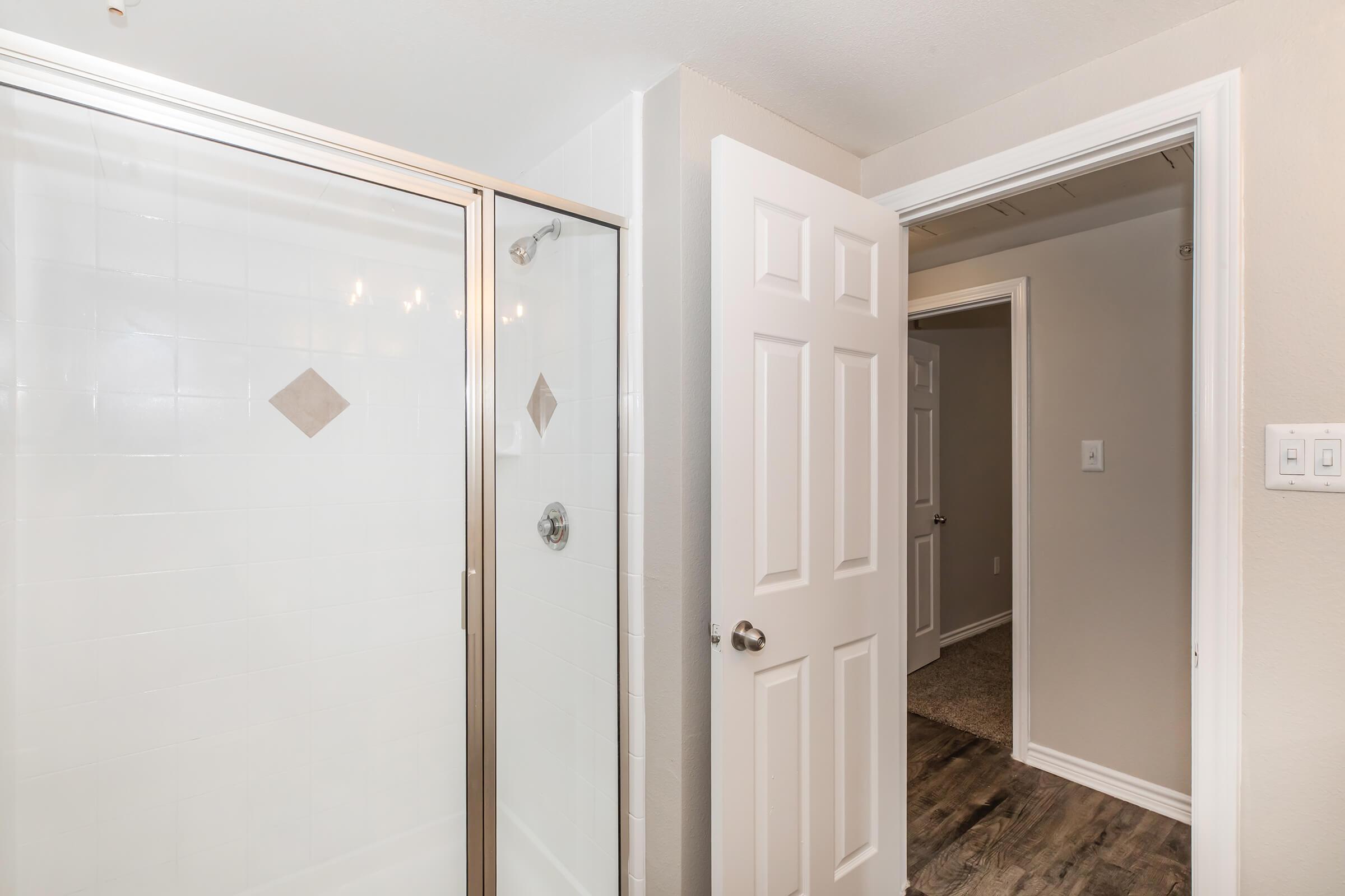 A clean bathroom interior featuring a glass shower enclosure with a tiled wall, light-colored walls, and a doorway leading to another room. The door is open, revealing a carpeted area beyond. The lighting is bright, creating a welcoming atmosphere.