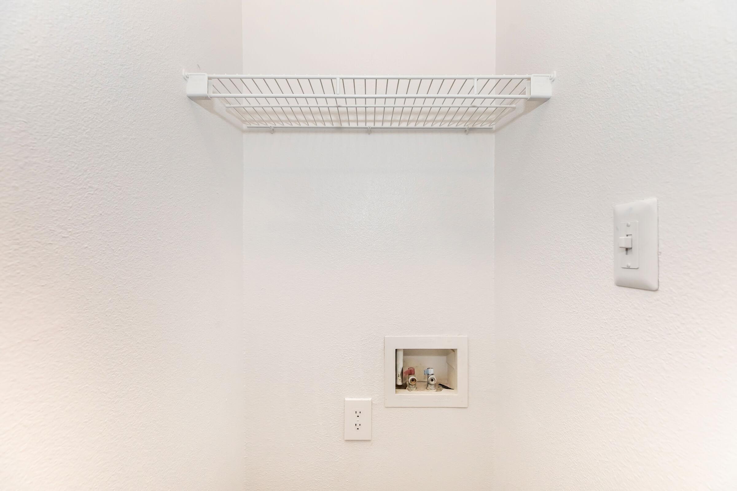 A small, empty closet featuring a white wire shelf mounted on the wall. There is a rectangular opening in the wall with an electrical outlet below it, and a small compartment in the wall that is open, displaying some items inside. The walls are painted white, contributing to a clean, minimalist look.