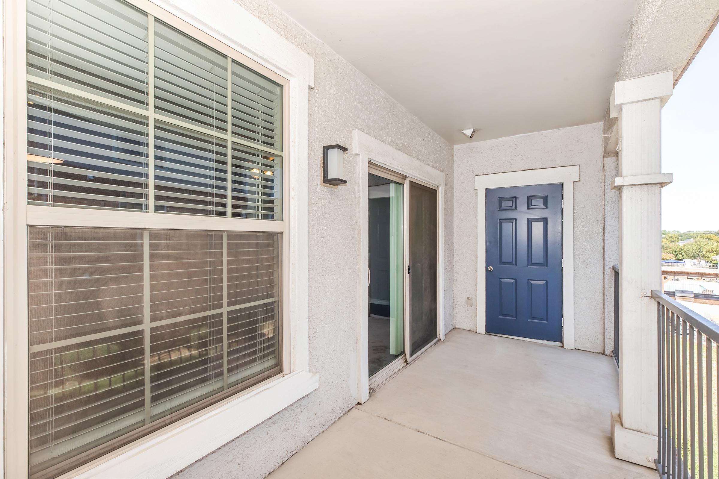 View of a small balcony area with a light-colored wall and large sliding glass door. The entrance features a blue door, and there are windows with horizontal blinds on one side. The floor is concrete, and the space is well-lit, suggesting a pleasant outdoor environment.
