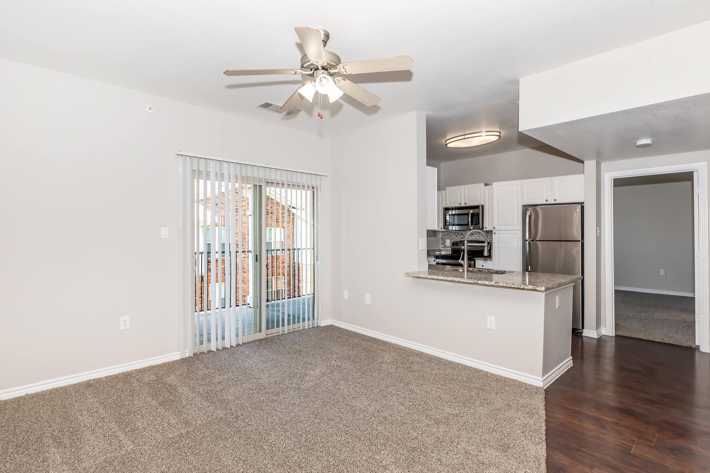 A spacious, well-lit living area with light beige walls and plush carpet. The room features a ceiling fan and has large sliding glass doors leading to a balcony. An open kitchen is visible with white cabinetry, stainless steel appliances, and a granite countertop. There is a hallway leading to another room in the background.