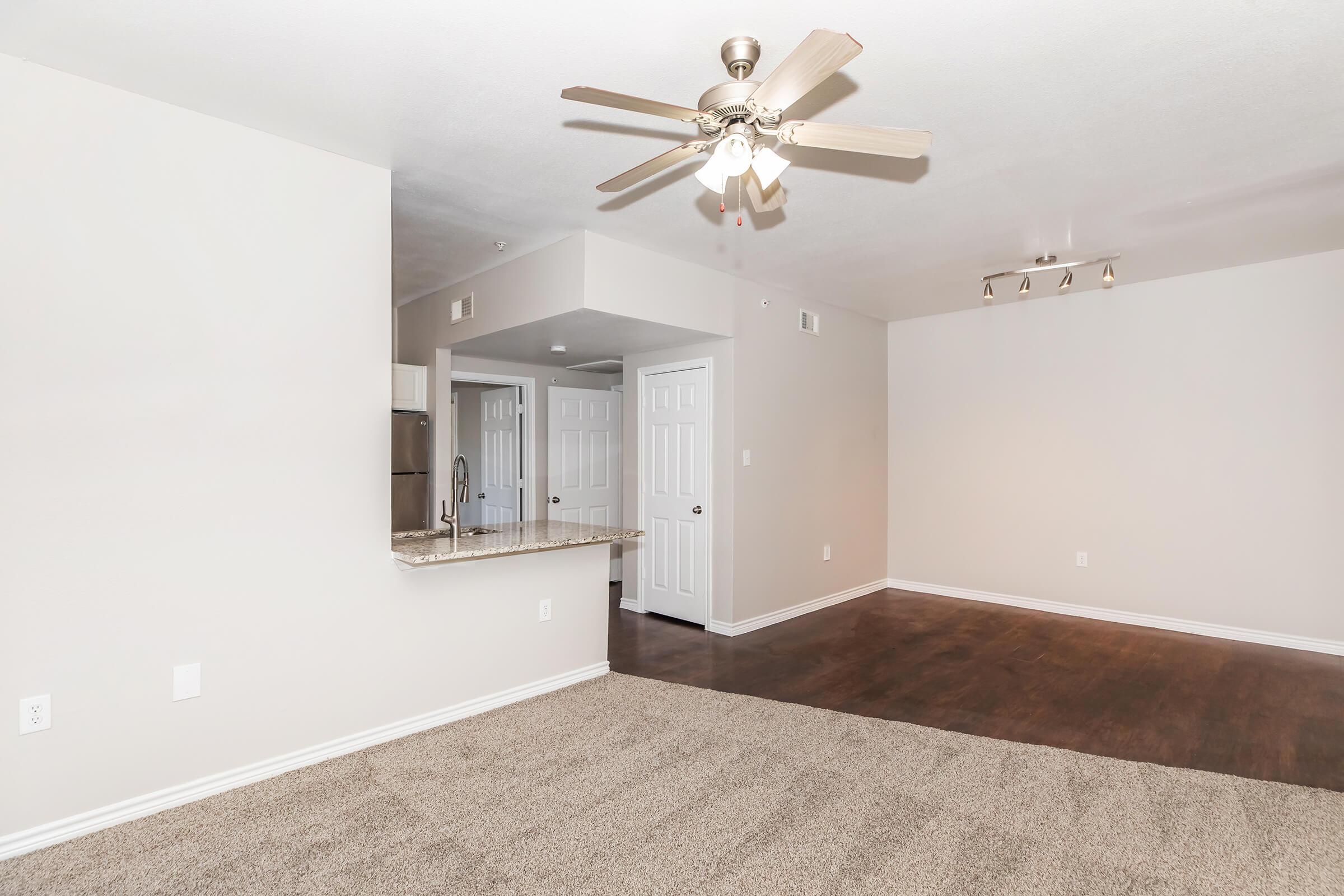 A spacious, well-lit living area featuring light beige walls and plush carpet. A ceiling fan hangs from the ceiling, and a kitchen area can be seen in the background with a countertop. The entrance to other rooms is visible through white doors on the left. The overall ambiance is modern and inviting.