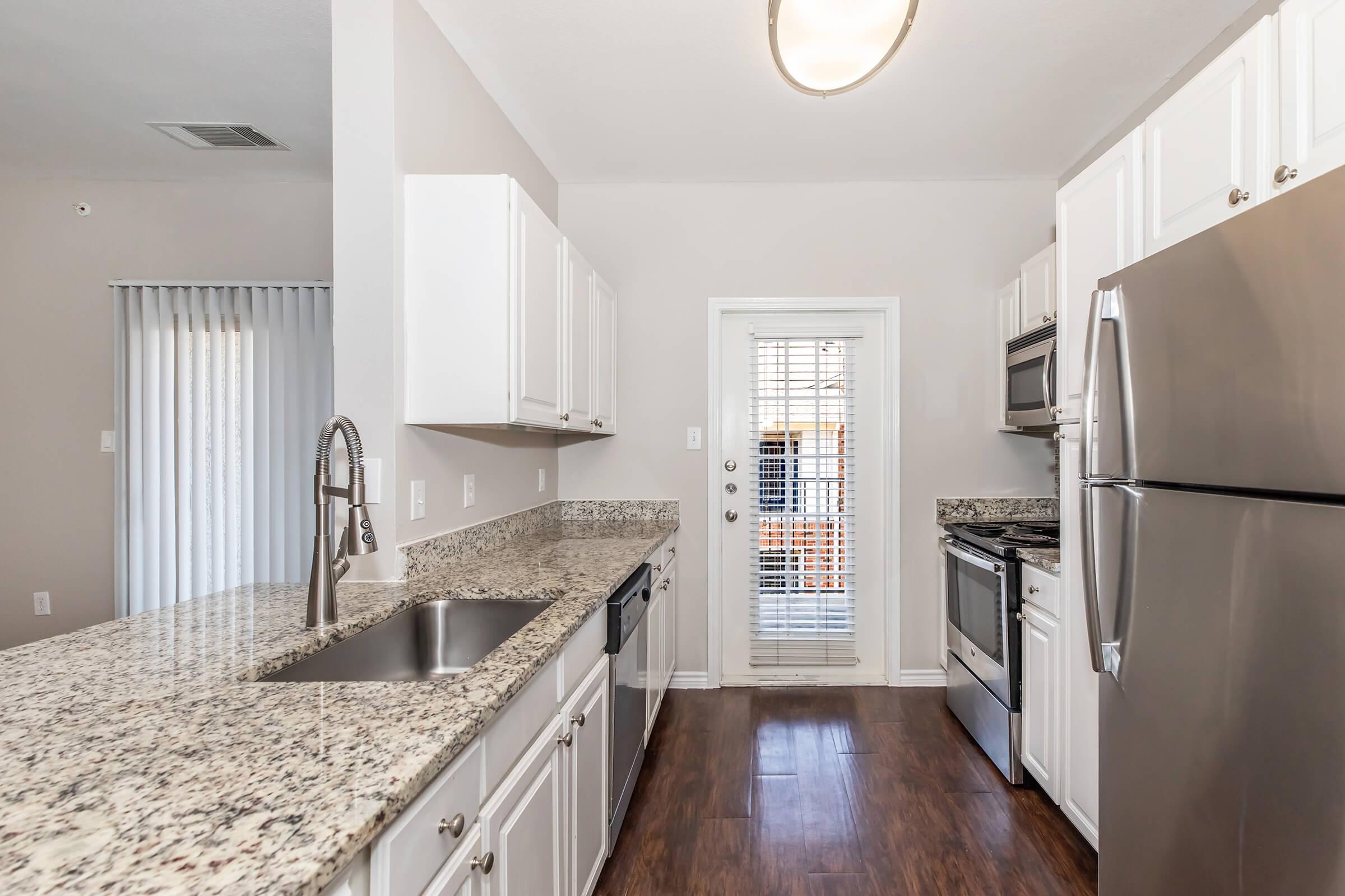 A modern kitchen featuring white cabinets, granite countertops, and stainless steel appliances. The space includes a double sink, a dishwasher, and an adjoining door leading to an outdoor area. Natural light filters through the window coverings, enhancing the welcoming atmosphere.
