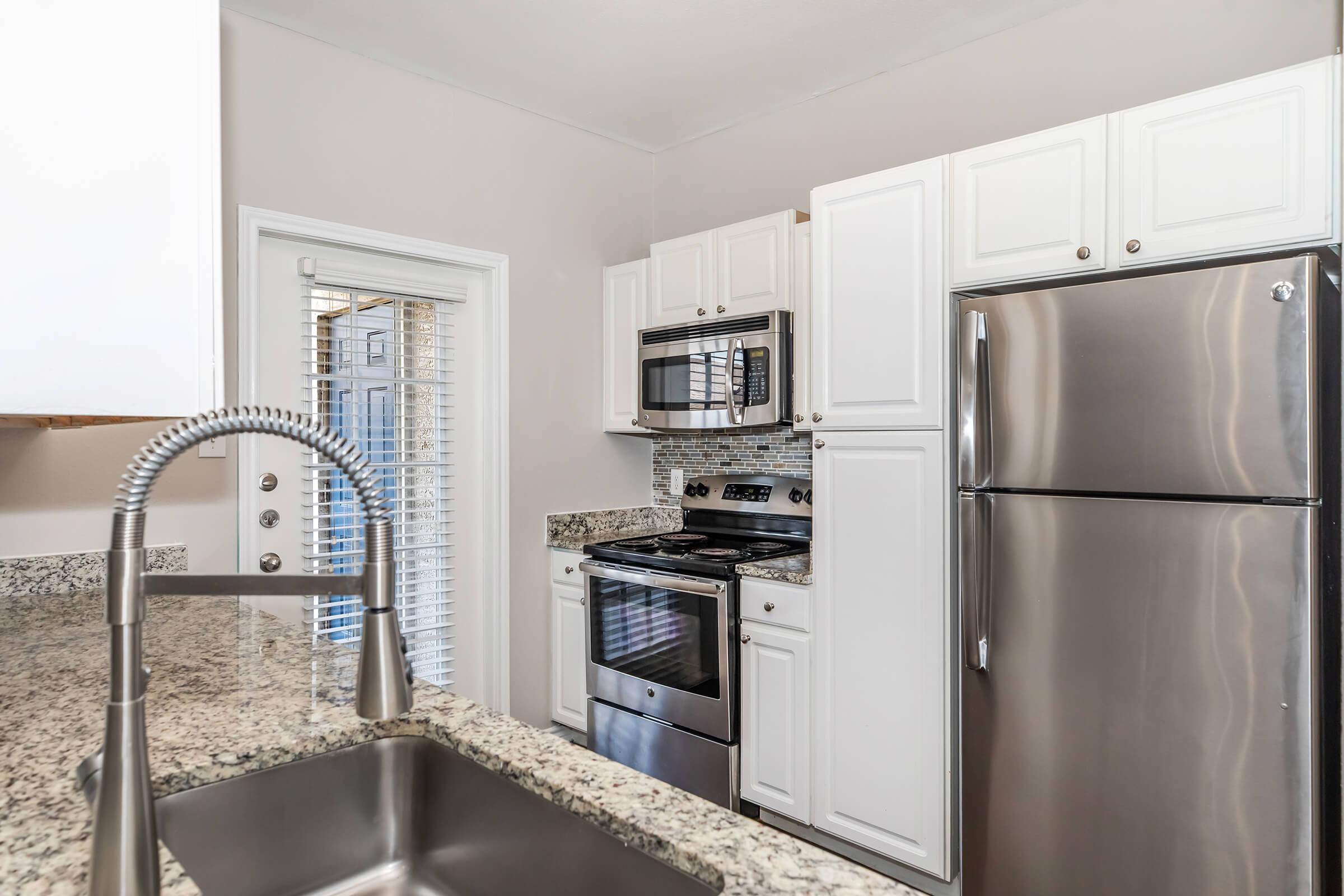 Modern kitchen featuring white cabinetry, stainless steel appliances, a granite countertop, and a double sink. The kitchen includes a built-in microwave, oven, and refrigerator, along with a view of the exterior through a sliding door with blinds. Bright, airy atmosphere with neutral walls.