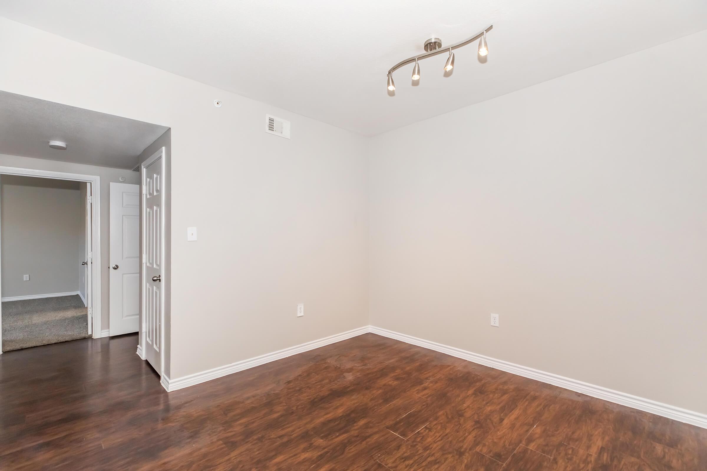Empty room featuring light brown wooden flooring and light gray walls. A ceiling light fixture with four bulbs is mounted above. The room has one open doorway leading to another area, and there are no furnishings or decorations visible, creating a spacious and minimalist appearance.