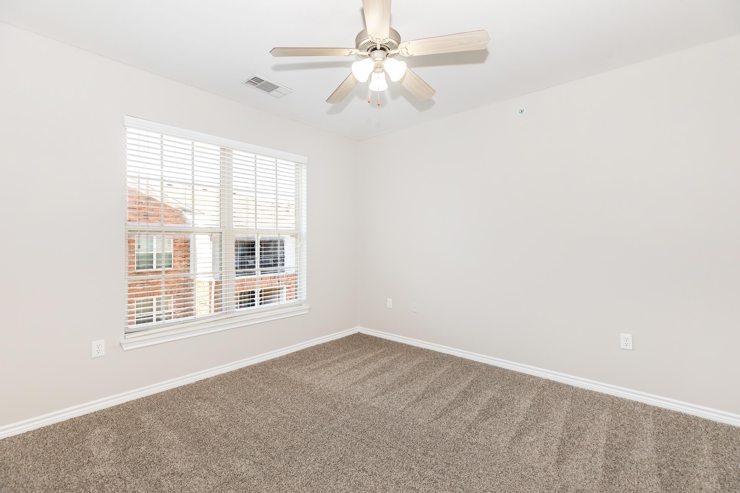 A spacious, empty room with light beige walls and plush carpet. The room features a ceiling fan with light fixtures and a large window with white blinds, allowing natural light to enter. The overall ambiance is bright and inviting, ideal for personal decoration or furnishing.
