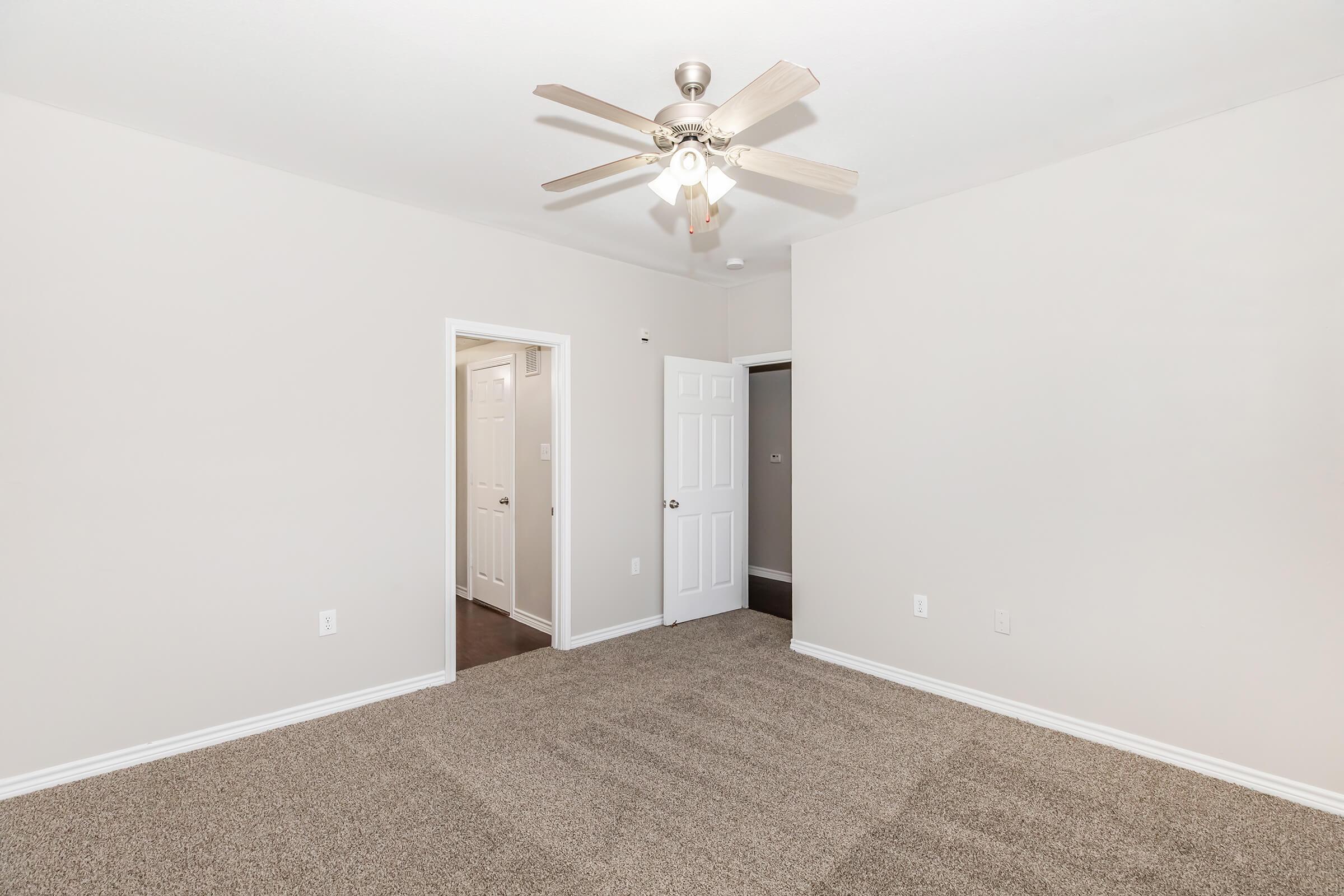 A light-colored room with beige carpet, featuring a ceiling fan with wooden blades. There are two doorways; one leads to a closet and the other to a hallway. The walls are plain and unadorned, creating a spacious and neutral atmosphere.