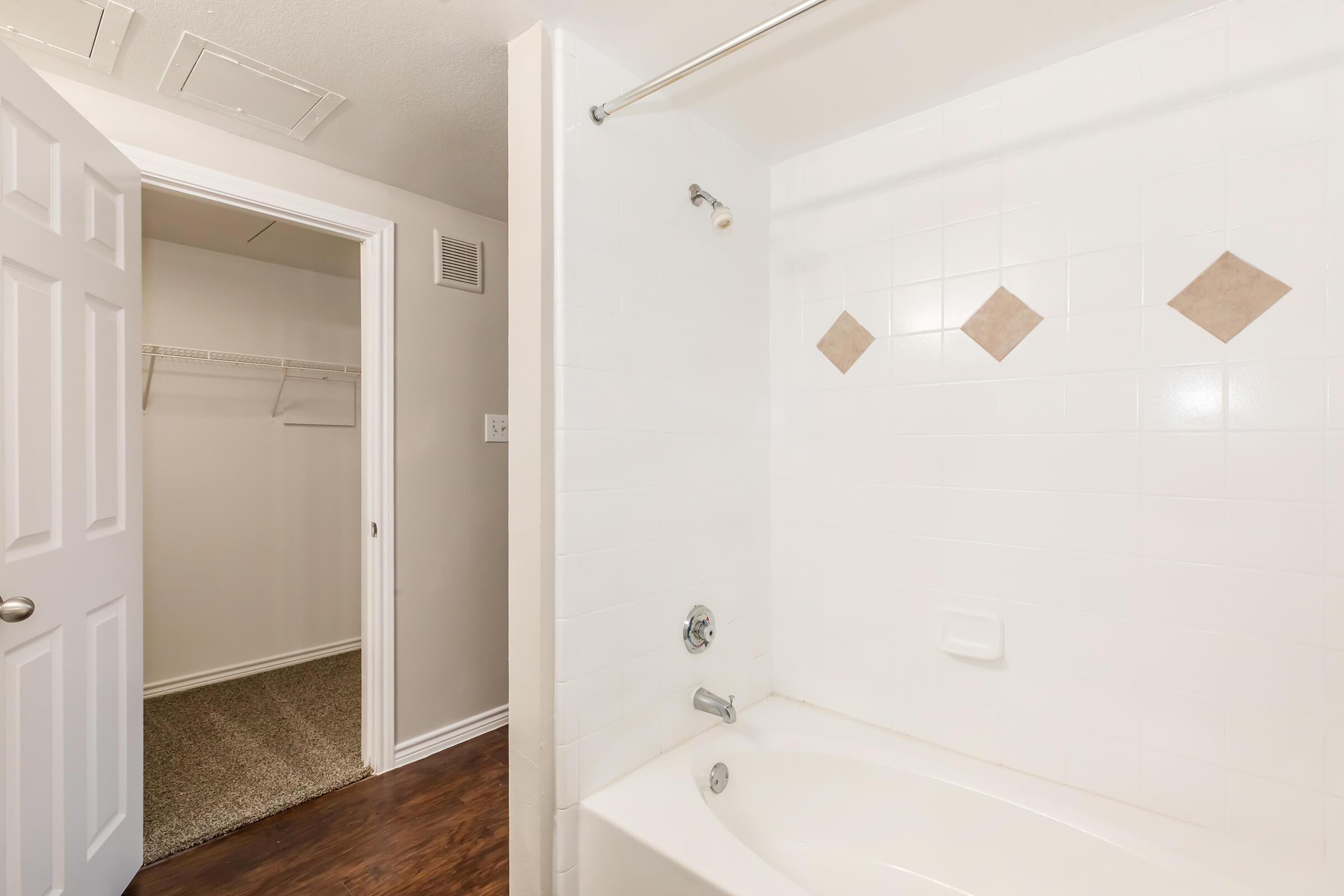 A clean bathroom featuring a white tiled bathtub with a showerhead, adjacent to a closet with wire shelving. The walls are painted in a light color, and the floor has dark wooden accents. The space appears well-maintained and organized, with a bright atmosphere.