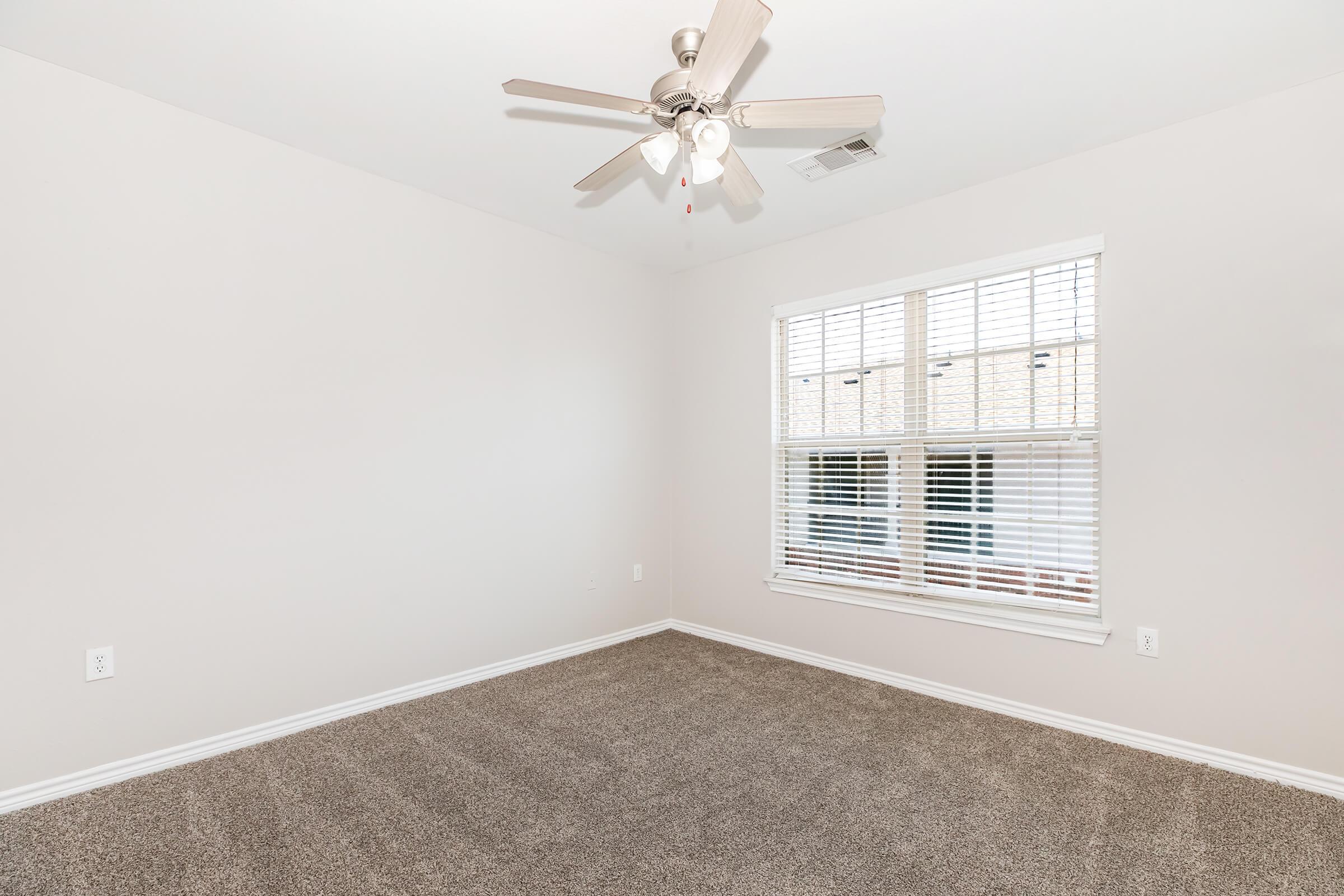A light-colored room with beige carpet, featuring a ceiling fan and a window with white blinds. The walls are painted in a soft neutral tone, creating a spacious and airy feel. Natural light enters through the window, illuminating the empty space, suitable for various uses.