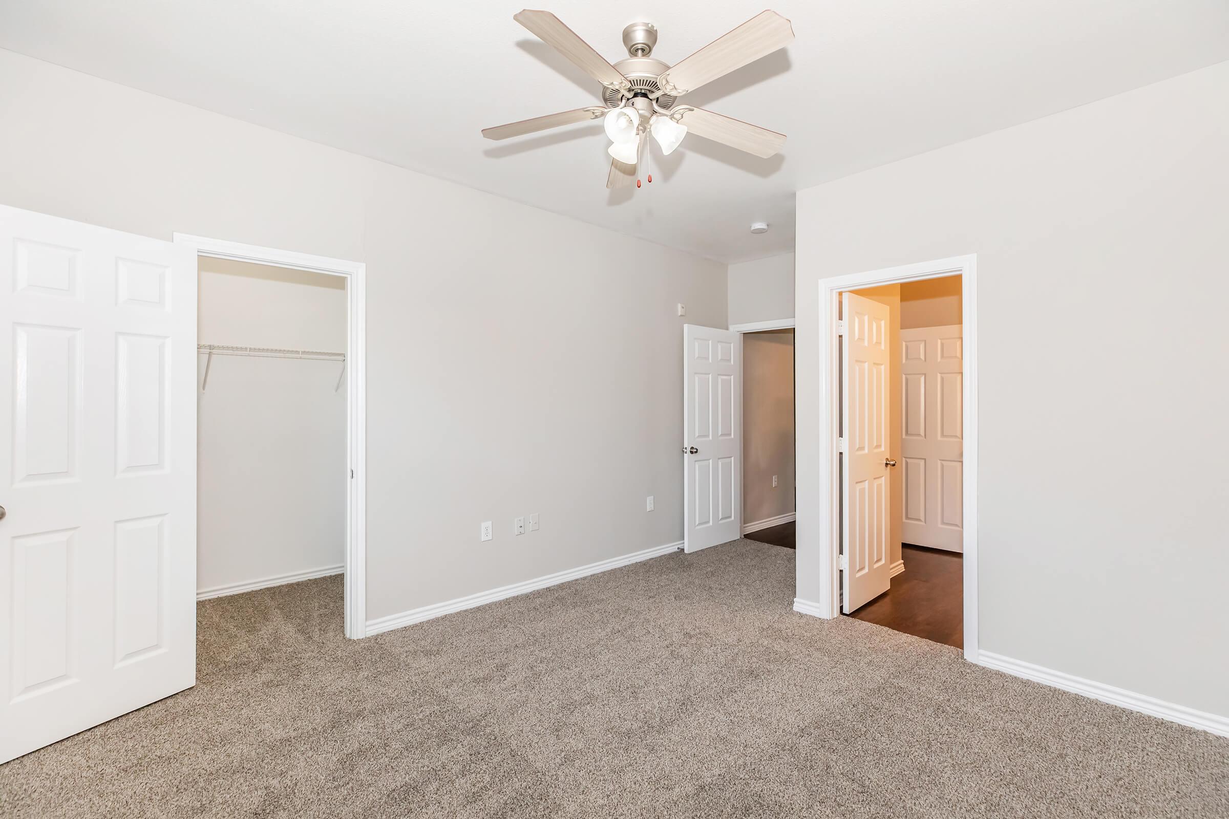 A well-lit empty bedroom featuring light gray walls, plush carpet, and a ceiling fan. Two doors lead to adjacent rooms, and one wall has an open closet space. The room is spacious and modern, with natural light illuminating the area.