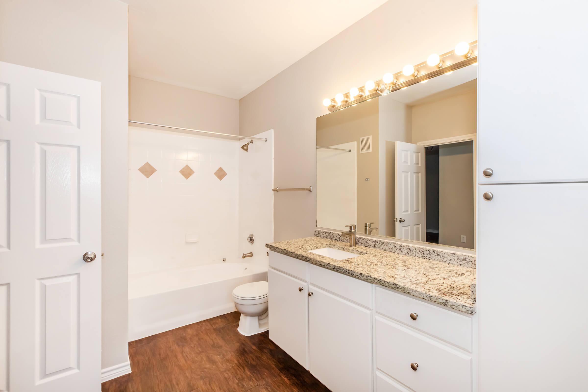 Bright and modern bathroom featuring a spacious layout with a white bathtub, toilet, and a sink with granite countertop. The room is well-lit by overhead lights and has light-colored walls, a large mirror, and wooden flooring. Storage cabinets are visible along the wall, providing additional space.