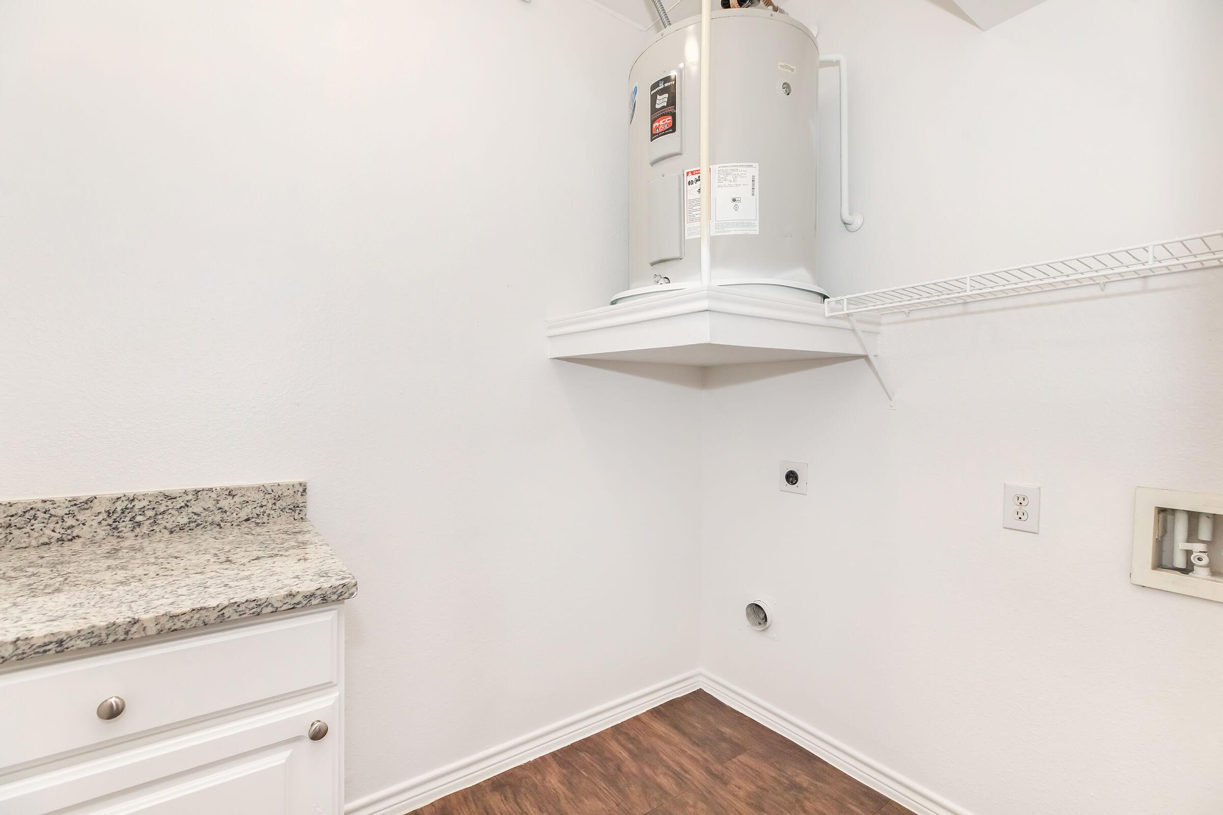 A clean laundry room featuring a white wall, a granite countertop, a water heater mounted on the wall, and a shelf above. The floor is covered with wood-like laminate. There's a washing machine hookup on the wall, and an electrical outlet is visible. The space is well-lit and organized.