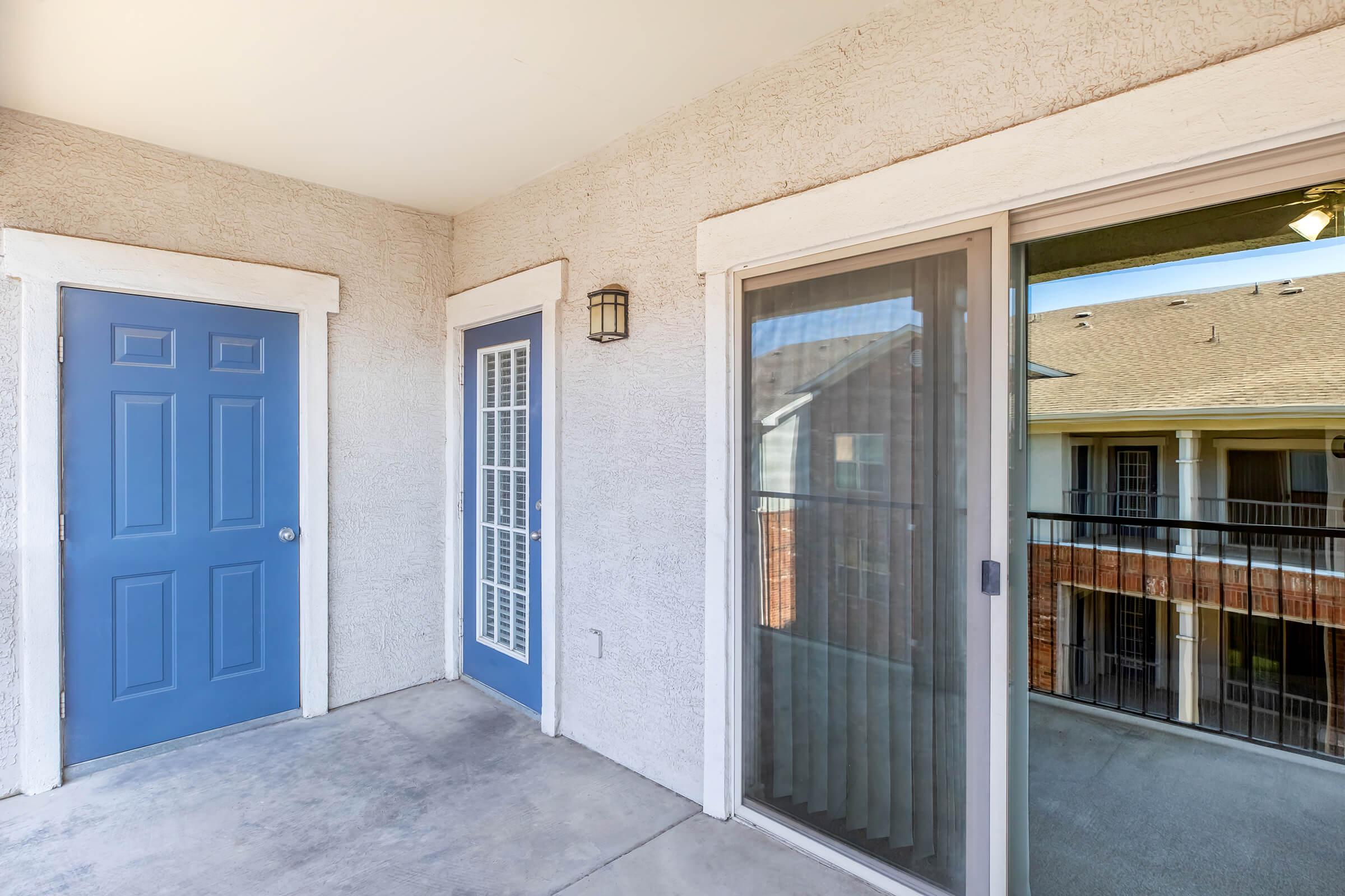 A balcony area of an apartment, featuring two doors: one blue, leading inside, and another with glass panels. The view shows a railing and surrounding architecture of the complex, with a well-maintained exterior and frosted glass. The scene is bright and inviting, highlighting a modern living space.