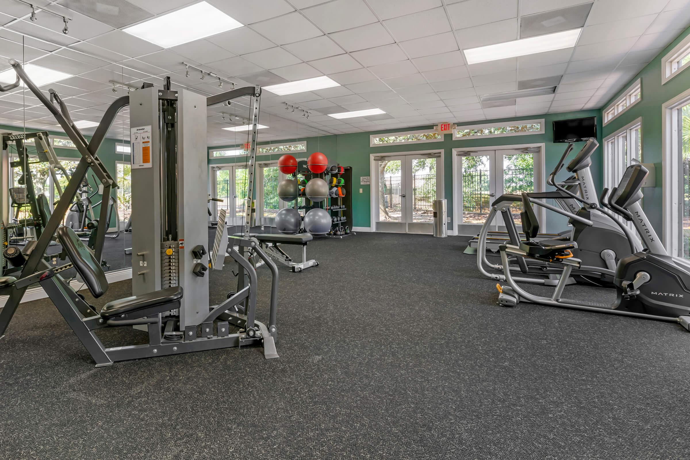 A modern gym interior featuring various exercise equipment including a multi-gym machine, an elliptical machine, and a collection of exercise balls arranged on colorful racks. The space is well-lit with large windows, offering a view of the outdoors and a clean, inviting atmosphere for workouts.