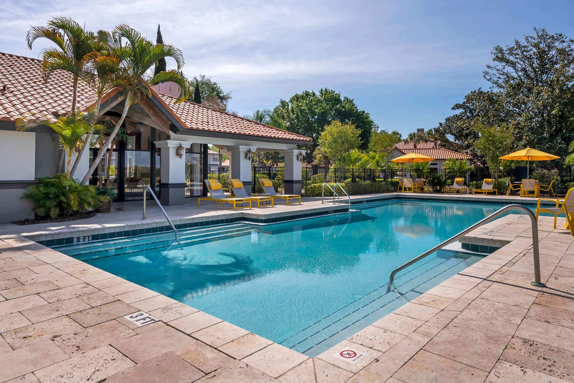 A clear blue swimming pool surrounded by patio tiles, lounge chairs, and tropical plants. The area includes yellow umbrellas and a building with a tiled roof in the background, under a bright blue sky. Ideal for relaxation and outdoor enjoyment.