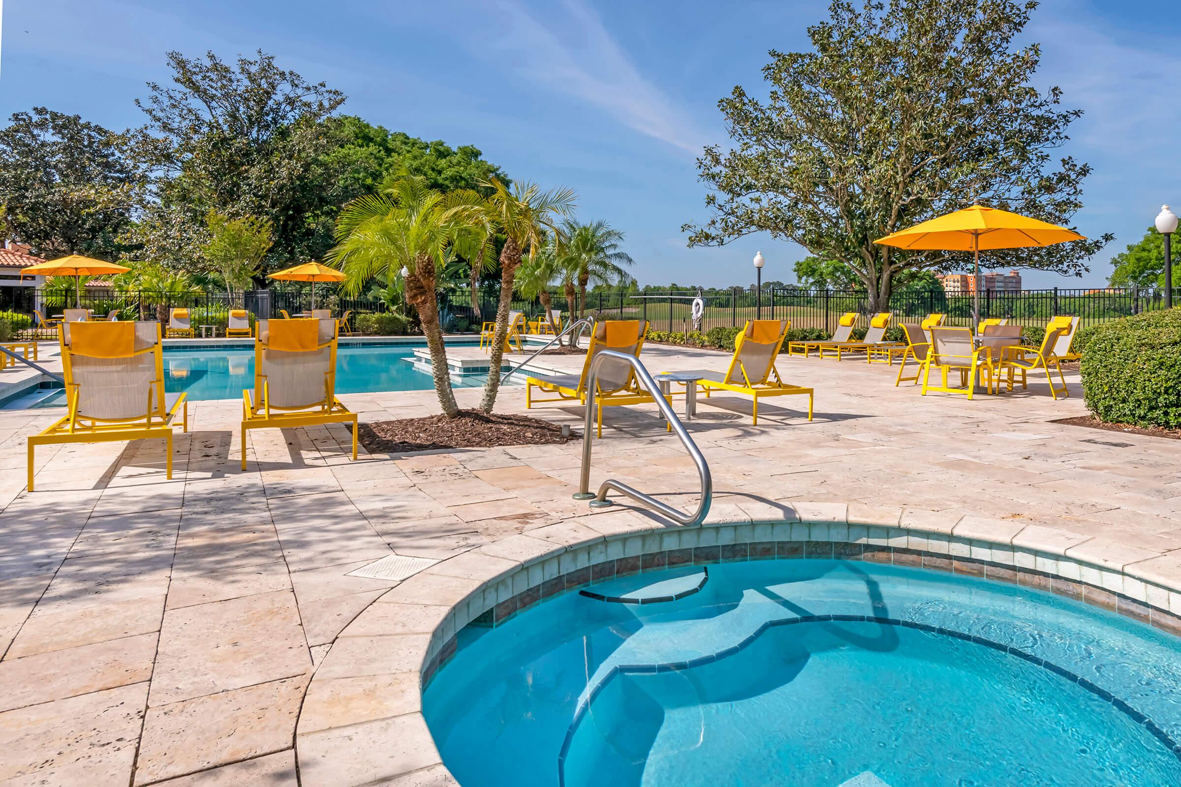 A serene outdoor swimming pool area featuring a sparkling blue pool and a hot tub. Surrounding the pool are yellow lounge chairs and umbrellas, set against a backdrop of lush trees and clear blue skies. The atmosphere is inviting and relaxing, perfect for a sunny day.