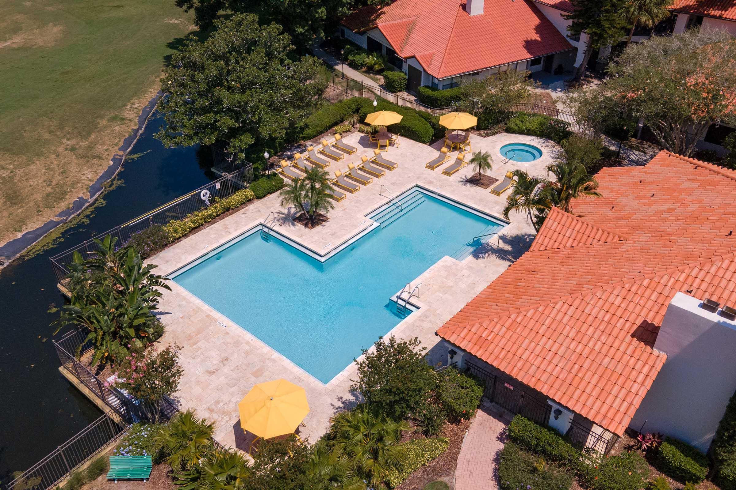 Aerial view of a swimming pool area featuring a large rectangular pool, a hot tub, and yellow umbrellas over lounge chairs. Surrounding the pool are lush green plants and trees, with a nearby water body and residential buildings with red-tiled roofs in the background.