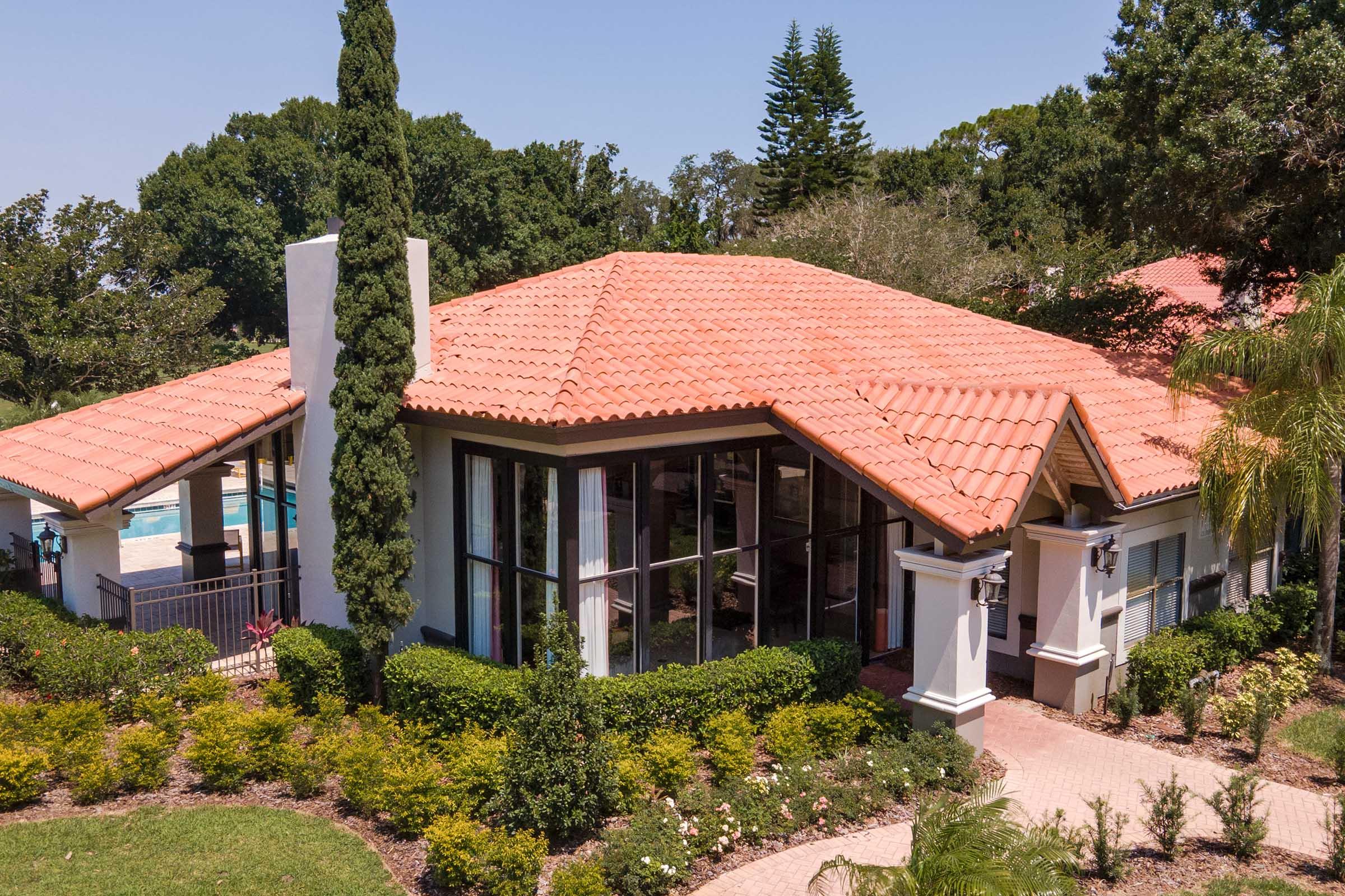 A modern single-story house with a red-tiled roof, surrounded by lush green landscaping, including palm trees and shrubs. The house features large windows and a spacious patio area, with a swimming pool visible in the background. The scene is set against a clear blue sky.