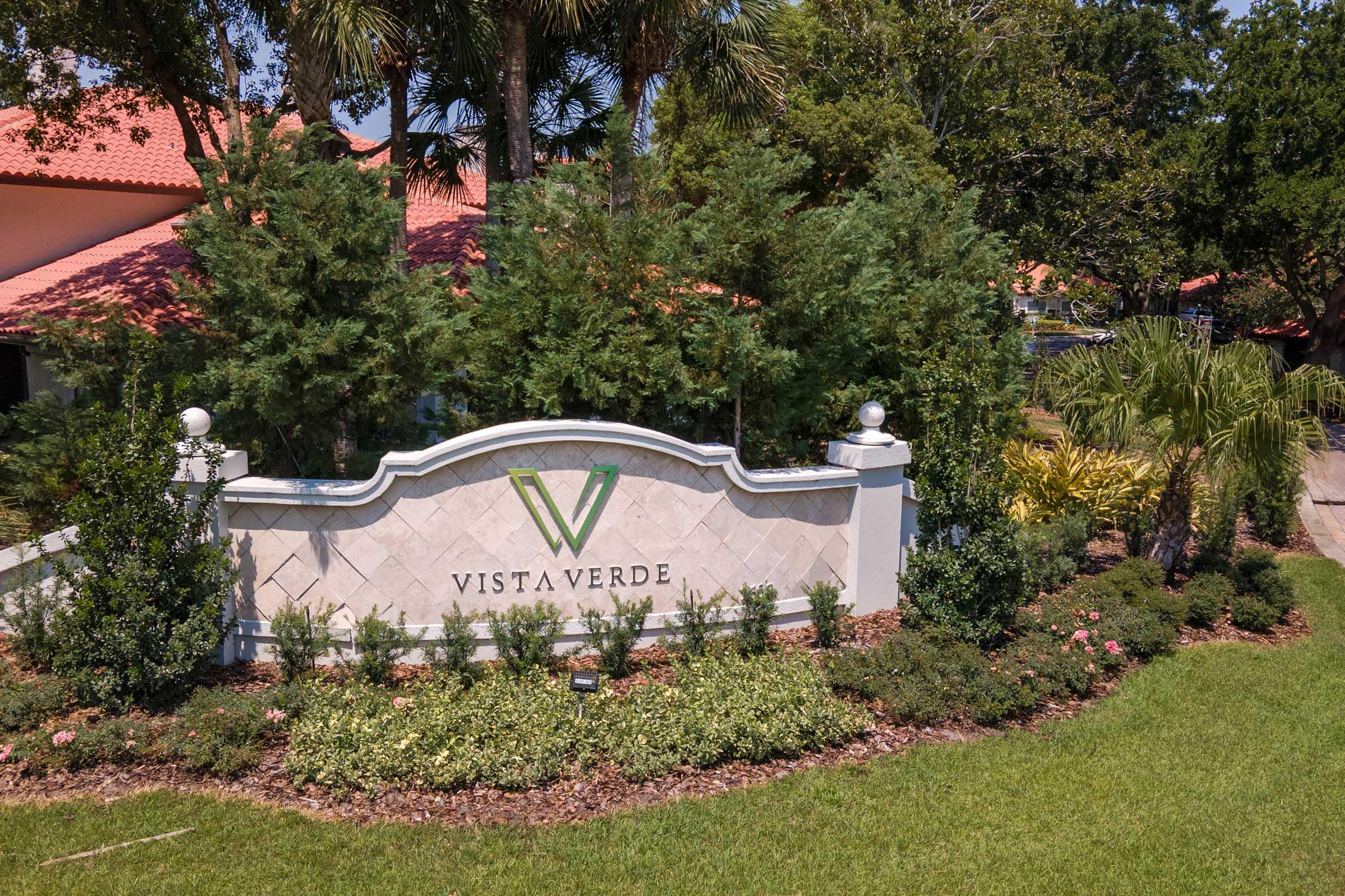 A well-manicured entrance sign of "Vista Verde" surrounded by lush greenery and decorative landscaping. The sign features a stylized “V” and is framed by a low stone wall, giving a welcoming impression to the community. Palm trees and plants enhance the picturesque setting.