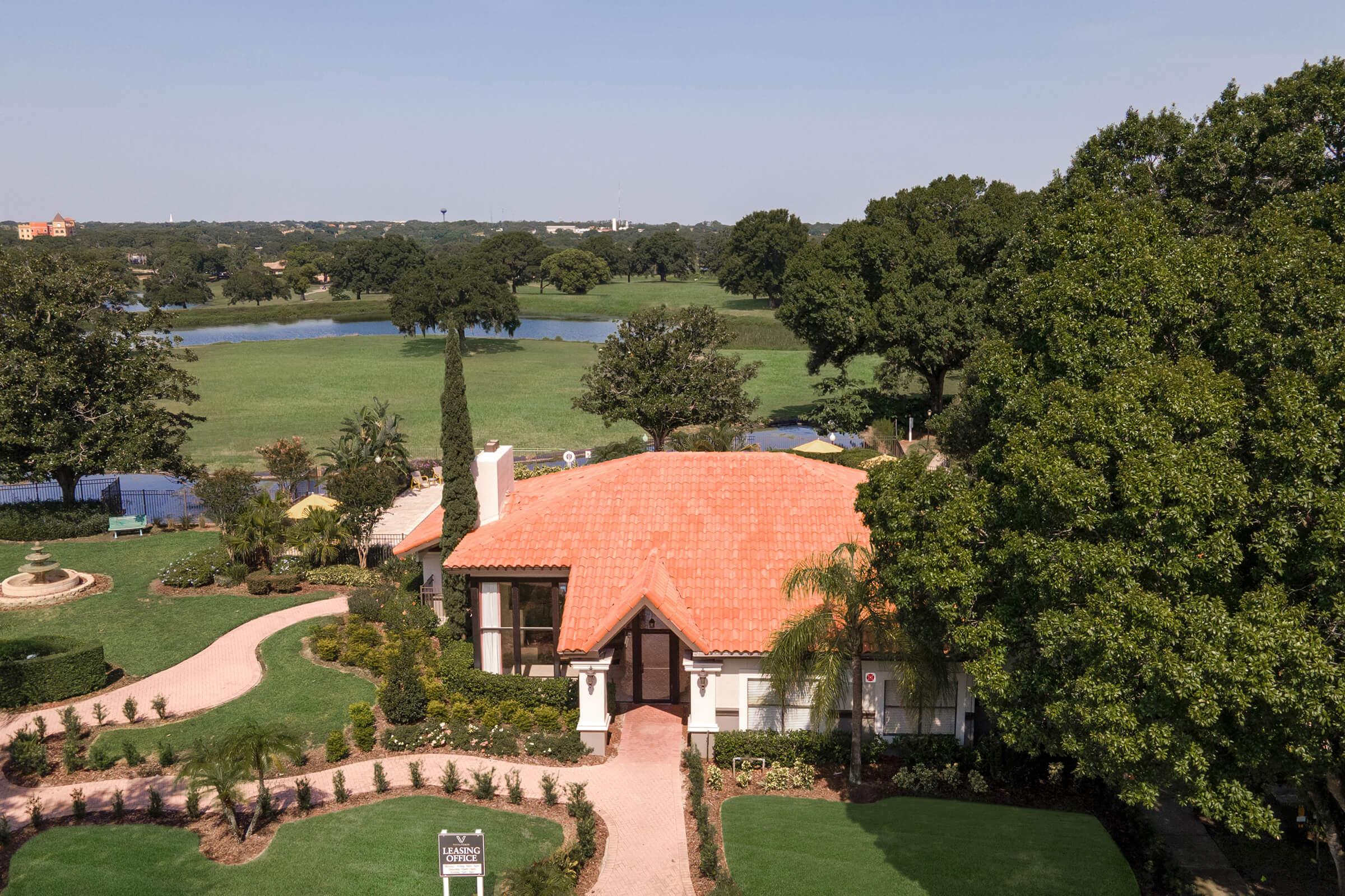 Aerial view of a charming house with a red-tiled roof, surrounded by lush green lawns, trees, and neatly arranged landscaping. A scenic body of water is visible in the background, with pathways and outdoor seating areas enhancing the serene atmosphere of the property.
