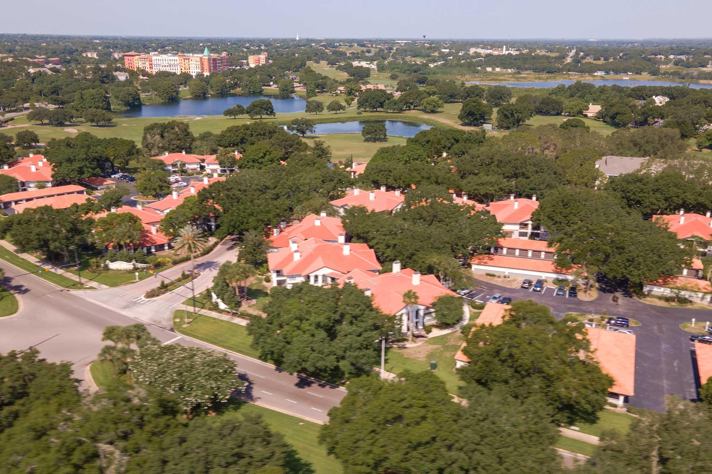 Aerial view of a suburban area featuring residential homes with red tile roofs, surrounded by lush greenery and trees. Multiple ponds and a larger building are visible in the background, indicating a well-planned community. The streets are lined with parked cars, adding to the residential atmosphere.