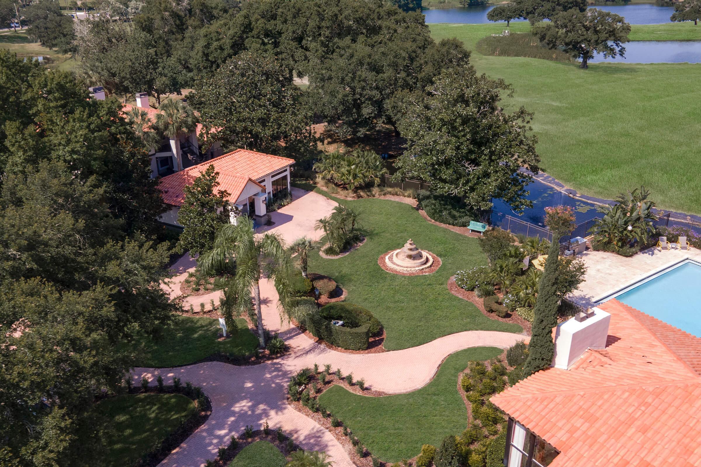 Aerial view of a landscaped garden featuring a circular pathway, neatly trimmed hedges, and a central fountain. There is a swimming pool on one side, surrounded by lush greenery, and an elegant house with a terracotta roof in the background, set against a scenic lake.