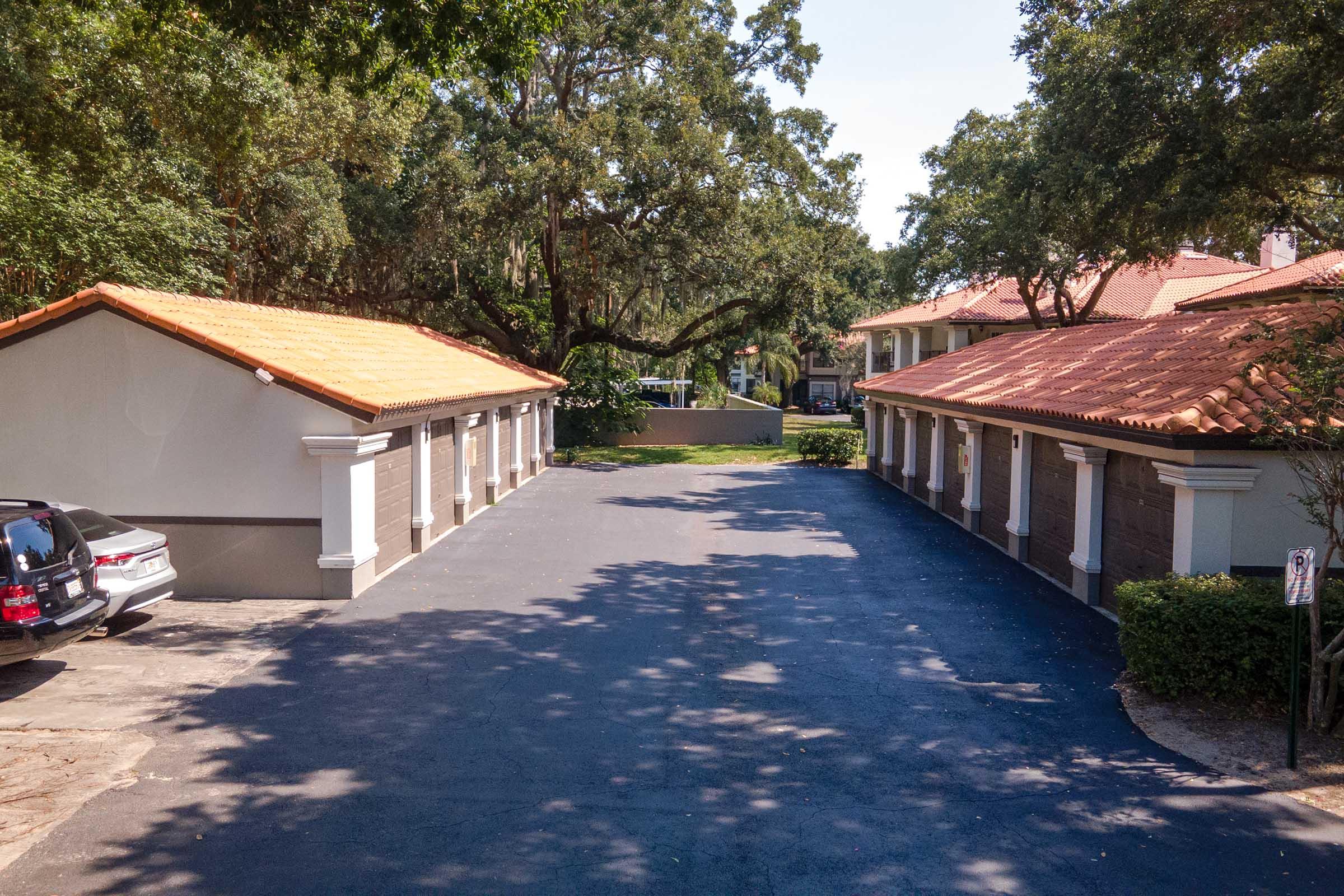 A view of a property with two buildings featuring tiled roofs on either side of a paved driveway. There are several trees providing shade, and parked cars are visible in the foreground. The scene is set on a sunny day, showcasing a well-maintained outdoor area.