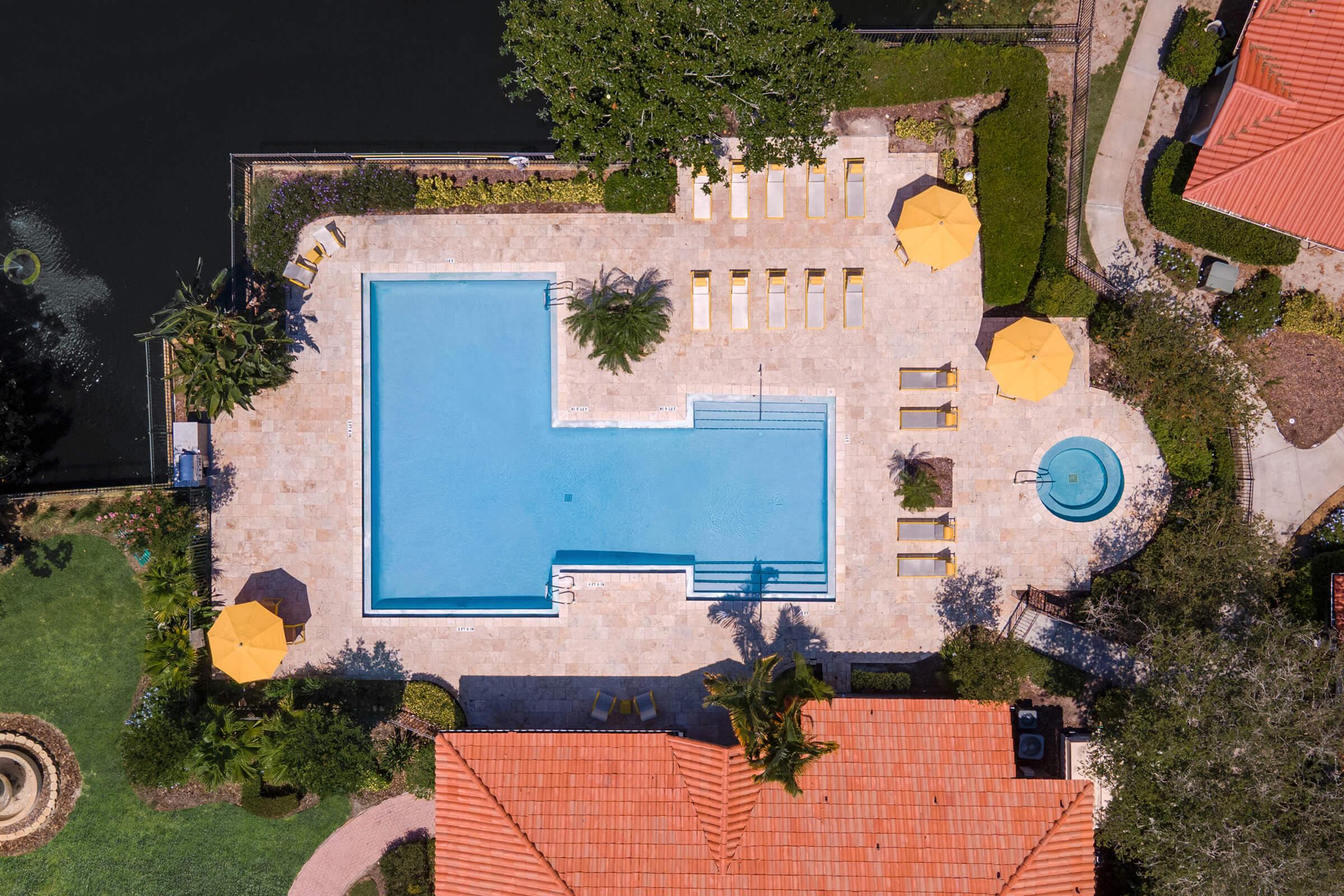 Aerial view of a rectangular swimming pool surrounded by lounge chairs and umbrellas, with a separate circular hot tub nearby. The pool area is bordered by lush greenery and houses with orange tiled roofs, overlooking a dark body of water.