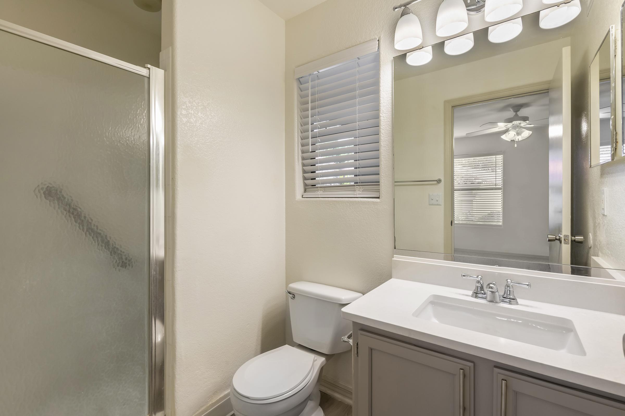 A clean and well-lit bathroom featuring a shower with a glass door, a toilet, and a double sink vanity with a mirror. Natural light streams in through a window with blinds, and there is a ceiling fan visible. The walls are painted in a light color, creating an open and airy feel.