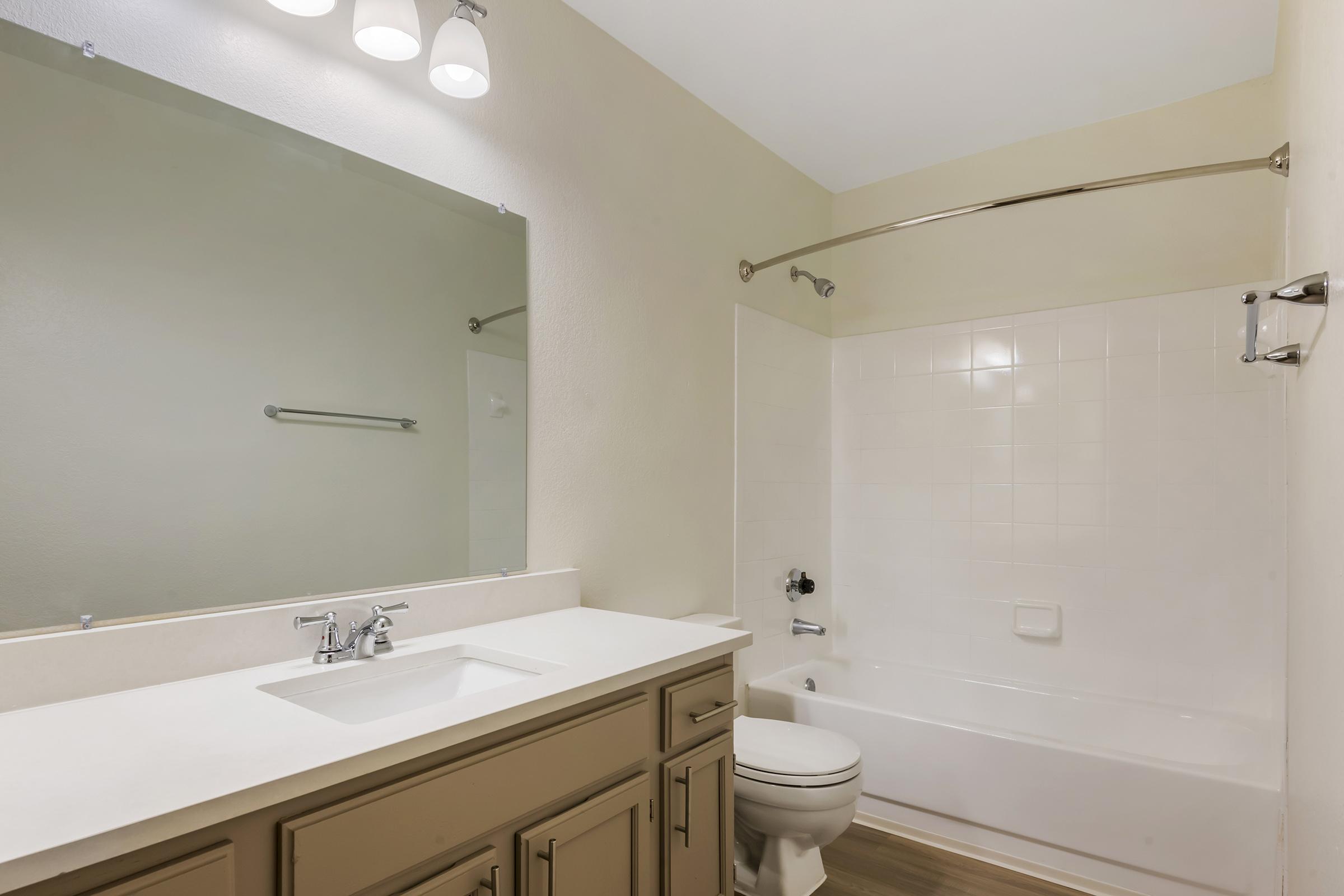 A clean, modern bathroom featuring a white bathtub with a shower, a mirror above a light-colored vanity with a sink, and neutral walls. Natural light is present, enhancing the spacious feel of the room.