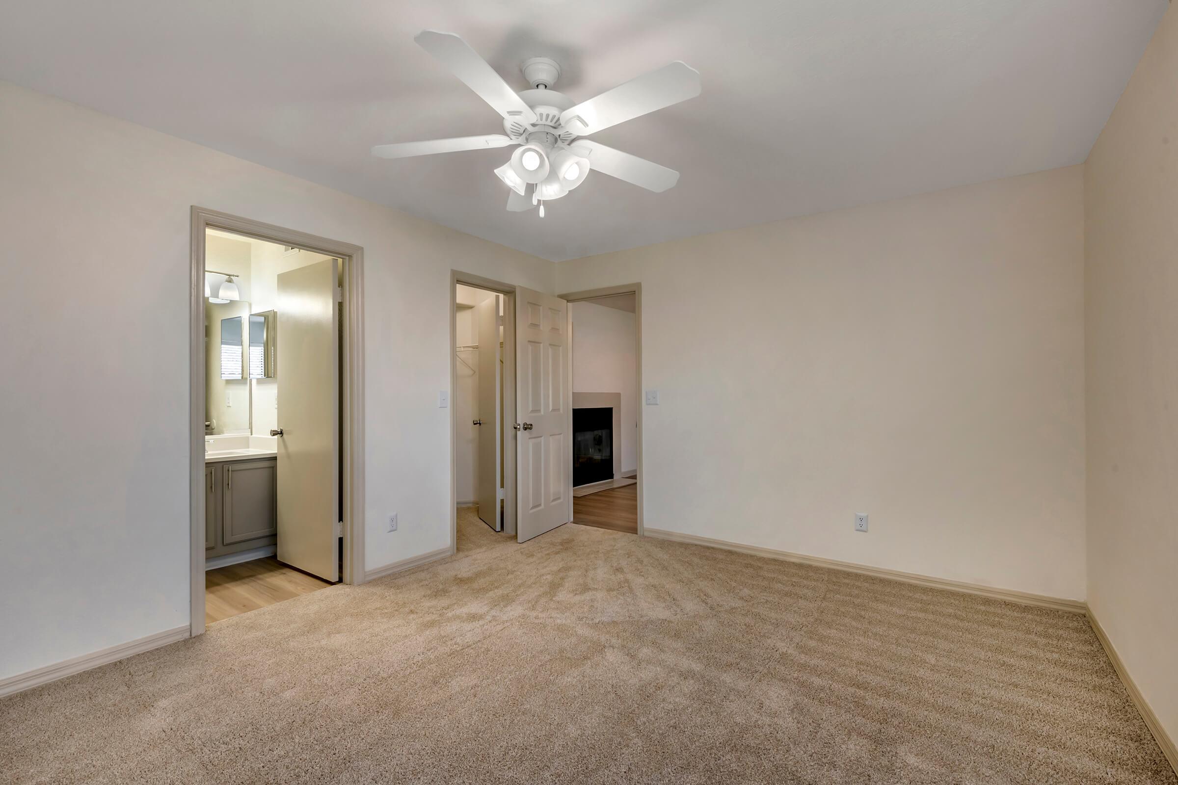 A well-lit, empty room featuring beige walls and carpet. A ceiling fan is mounted above. There are two doorways: one leads to a bathroom with a mirror and the other to a living space. The room is spacious and tidy, with natural light coming from the hallway.