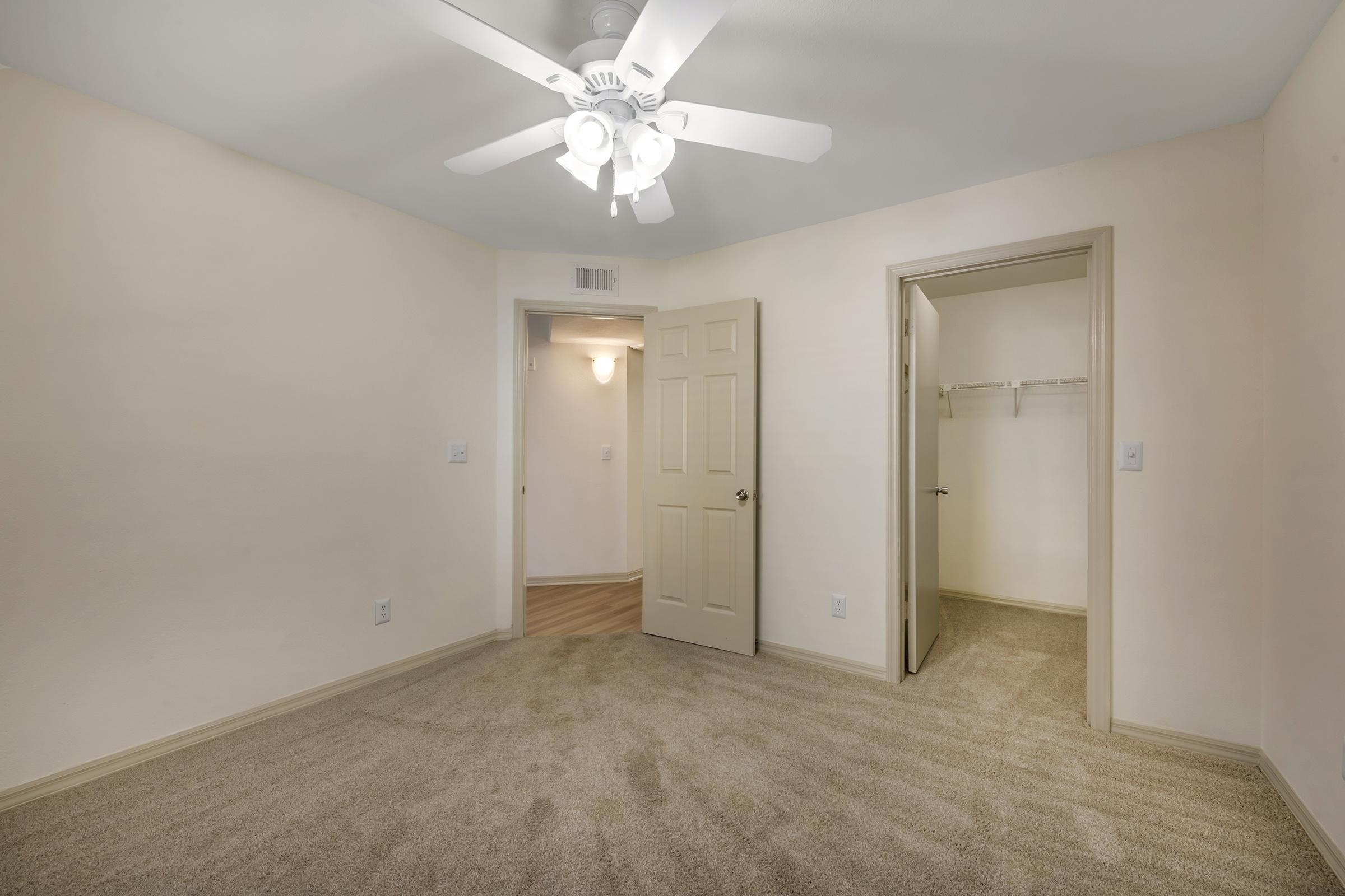 A light-colored, unfurnished bedroom featuring beige walls and a ceiling fan. There is a closed door leading to a closet on one side and an open door to another room on the other side. The carpet is a soft beige color, providing a clean and inviting atmosphere.