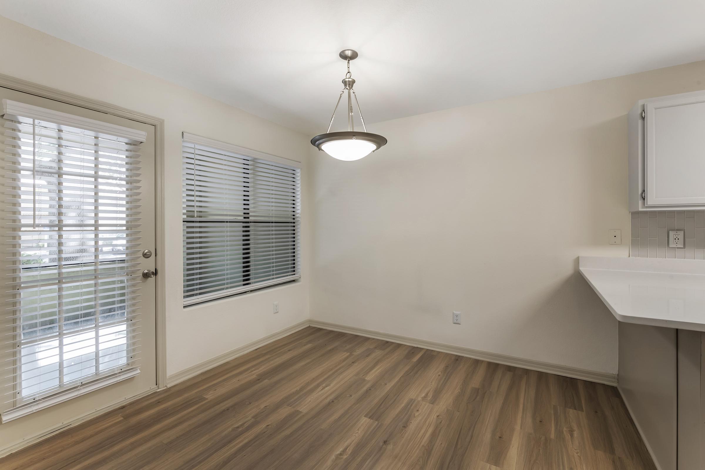 An empty room with a large window featuring blinds, a modern light fixture hanging from the ceiling, and light-colored walls. The flooring is made of wood, and there's a door leading to an outdoor area. A kitchen countertop is visible in the corner, suggesting a residential space.