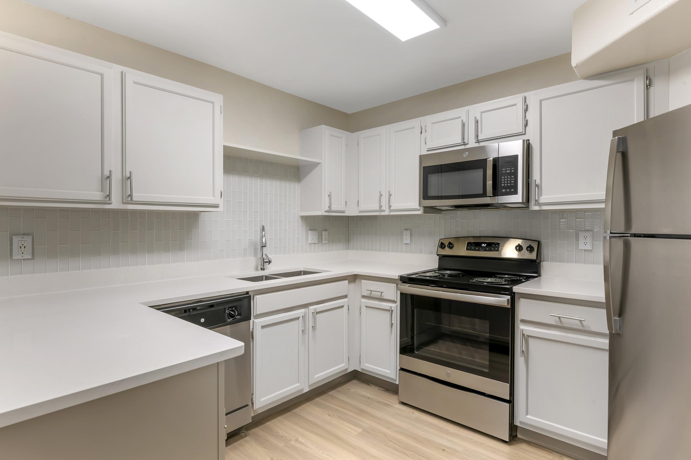 Modern kitchen featuring white cabinetry, stainless steel appliances including a microwave and oven, a sink with a modern faucet, and a light-colored countertop. The layout is open and spacious with ample natural light and a neutral color palette.