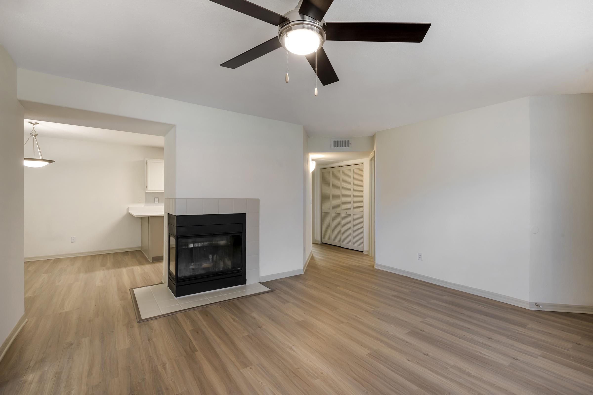 A spacious living area featuring light-colored walls and hardwood floors, with a ceiling fan for ventilation. There is a modern fireplace in the corner and a doorway leading to another room. Natural light fills the space, creating a warm and inviting atmosphere. A dining area is visible in the background.