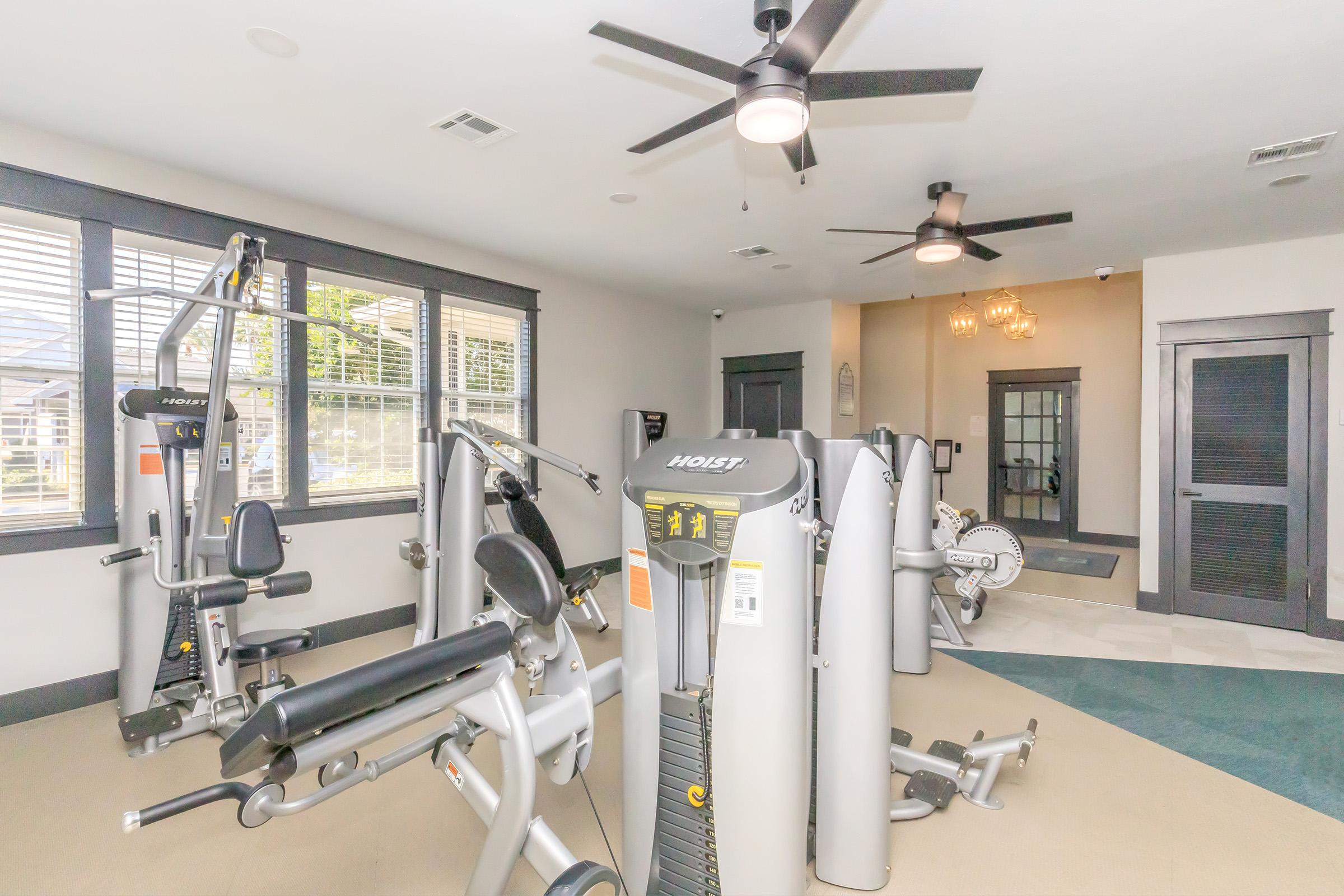 A well-lit fitness room featuring various exercise machines, including a multi-station weight machine and cardio equipment. The space has large windows, ceiling fans, and modern decor, creating a comfortable environment for workouts.