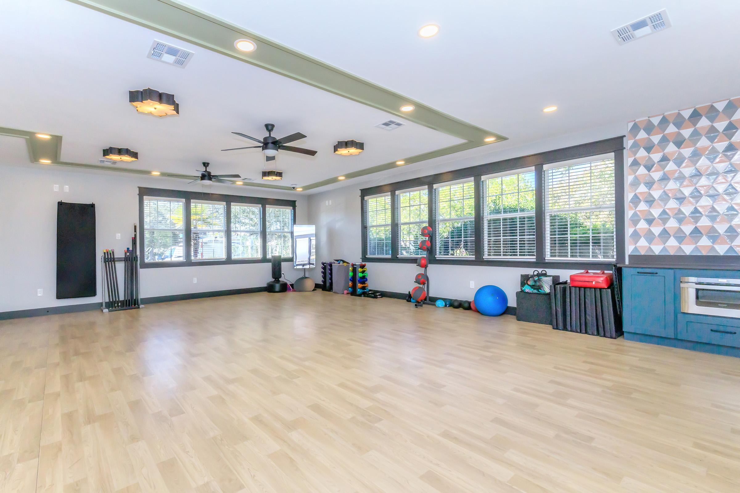 A spacious room featuring wooden flooring, large windows with blinds, and modern lighting. The room is equipped with exercise equipment including weights, a stability ball, and a mirrored wall, creating an inviting atmosphere for fitness activities. The decor includes a geometric patterned accent wall.