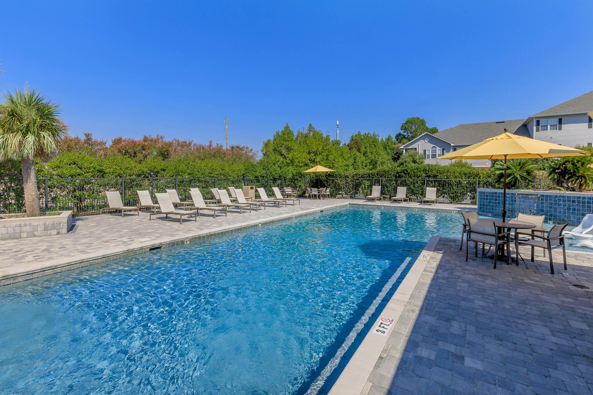 A clear blue swimming pool surrounded by lounge chairs and umbrellas, set in a landscaped area with greenery in the background. The sky is bright blue, and there are tables and chairs near the pool, creating a relaxing outdoor space.
