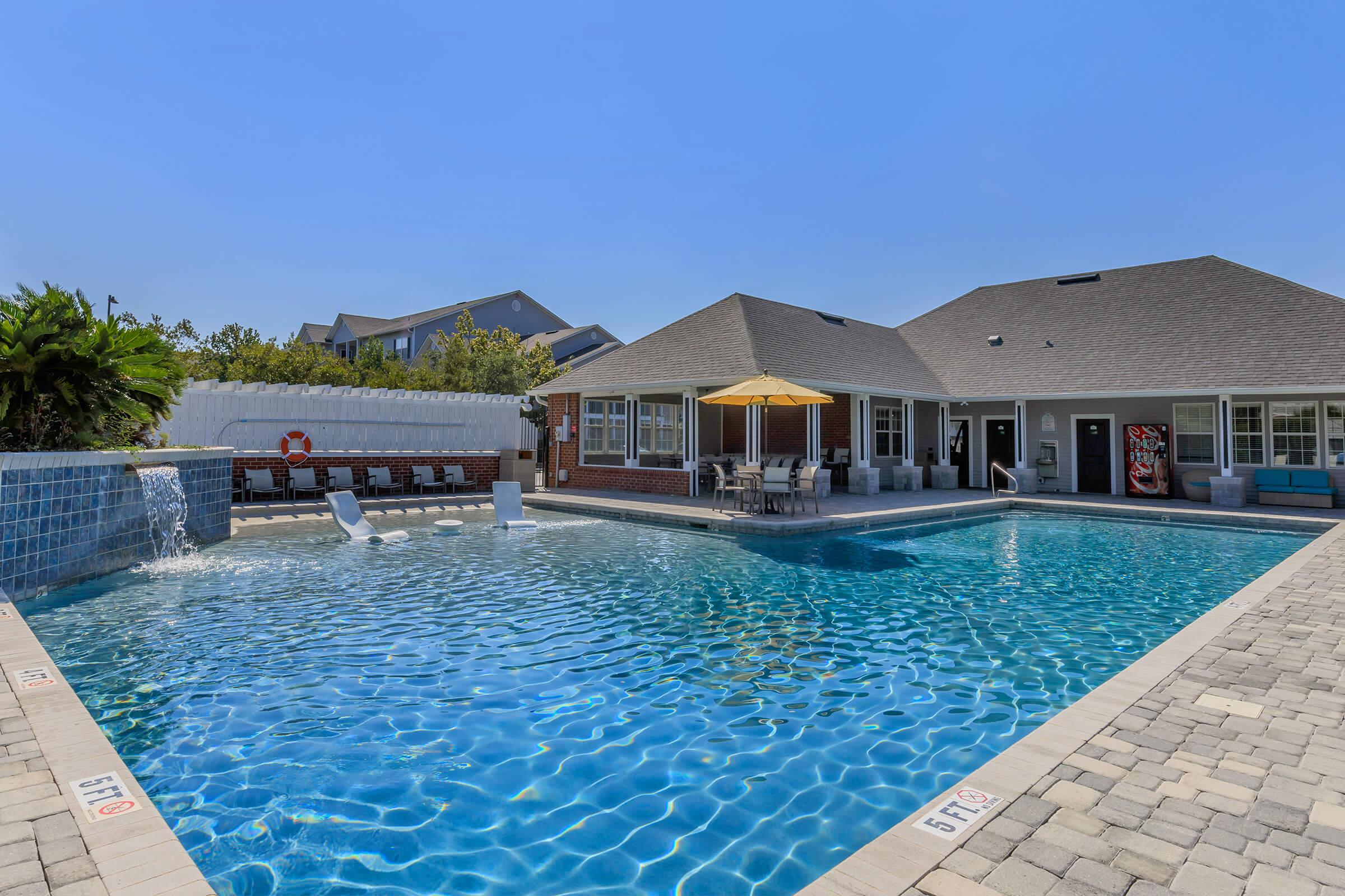 A sparkling blue pool surrounded by a spacious patio. The area features lounge chairs, an umbrella, and a shaded seating space. A fence and landscaped greenery provide privacy, while a nearby house with multiple windows adds to the setting. Clear blue sky overhead enhances the inviting atmosphere.